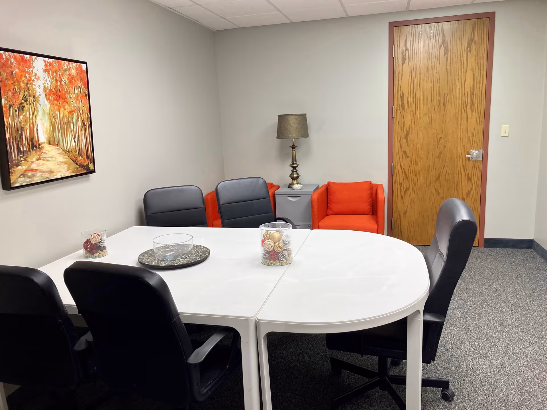 A round white dining table with black chairs, orange armchairs, and a lamp in a lounge.