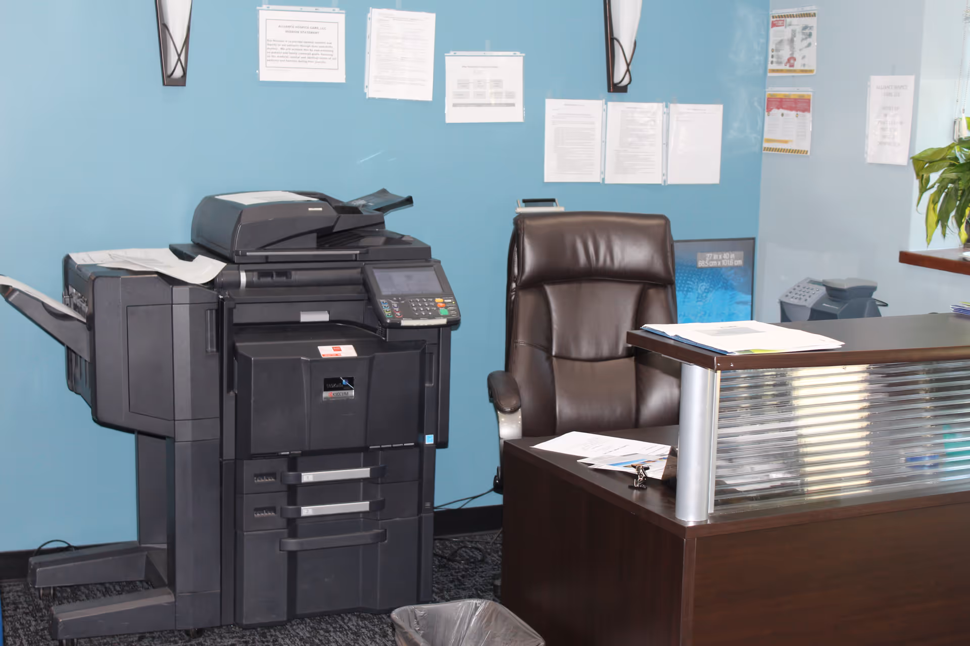 Office reception area with a large printer, desk, and brown chair against blue walls.