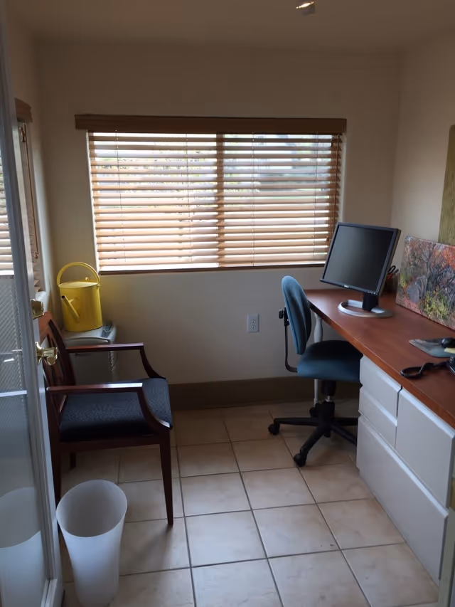 Small office nook with desk, computer, chair, blinds, and window in a senior living facility.