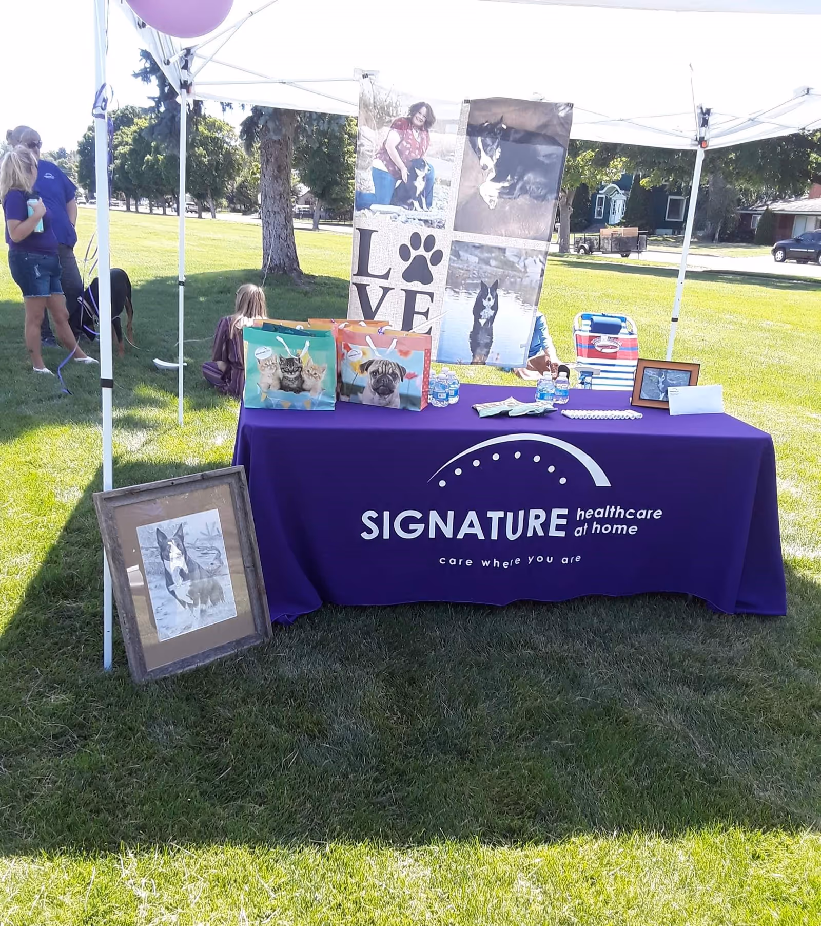 Outdoor tent booth with a purple Signature HealthCare at Home table and photo posters.