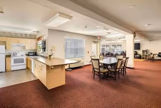 Open-plan common area with kitchen island, dining table, and seating in a senior living facility.