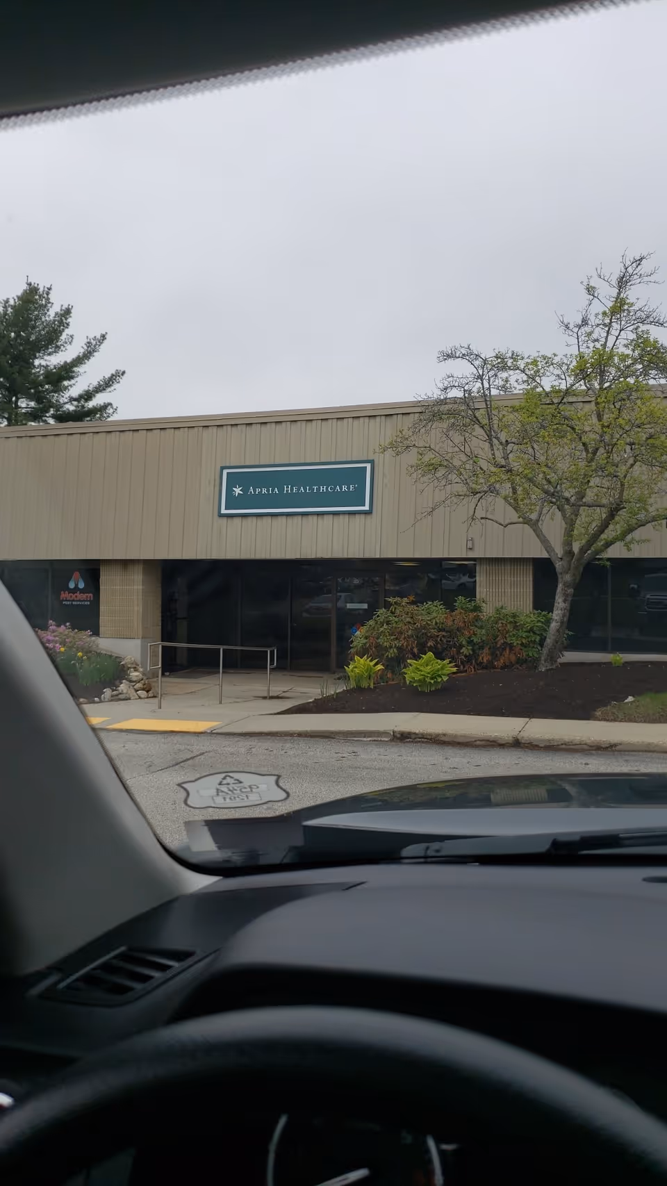View from inside a car of Apria Healthcare building front with sign and landscaping.