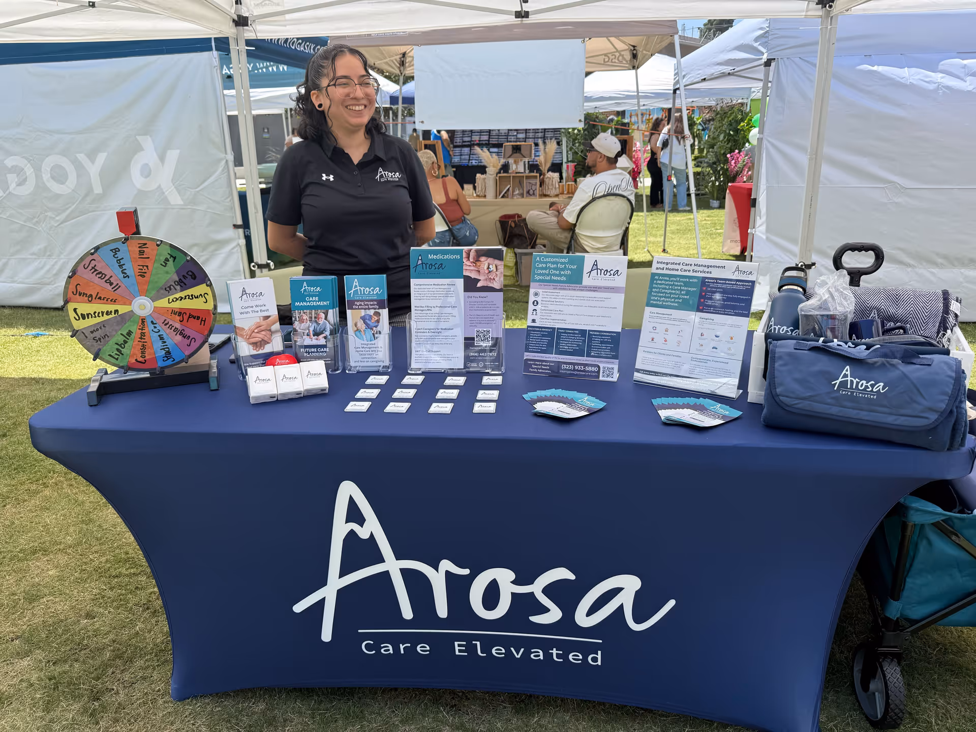 Arosa booth at an outdoor event with a table of brochures and a smiling staff member.