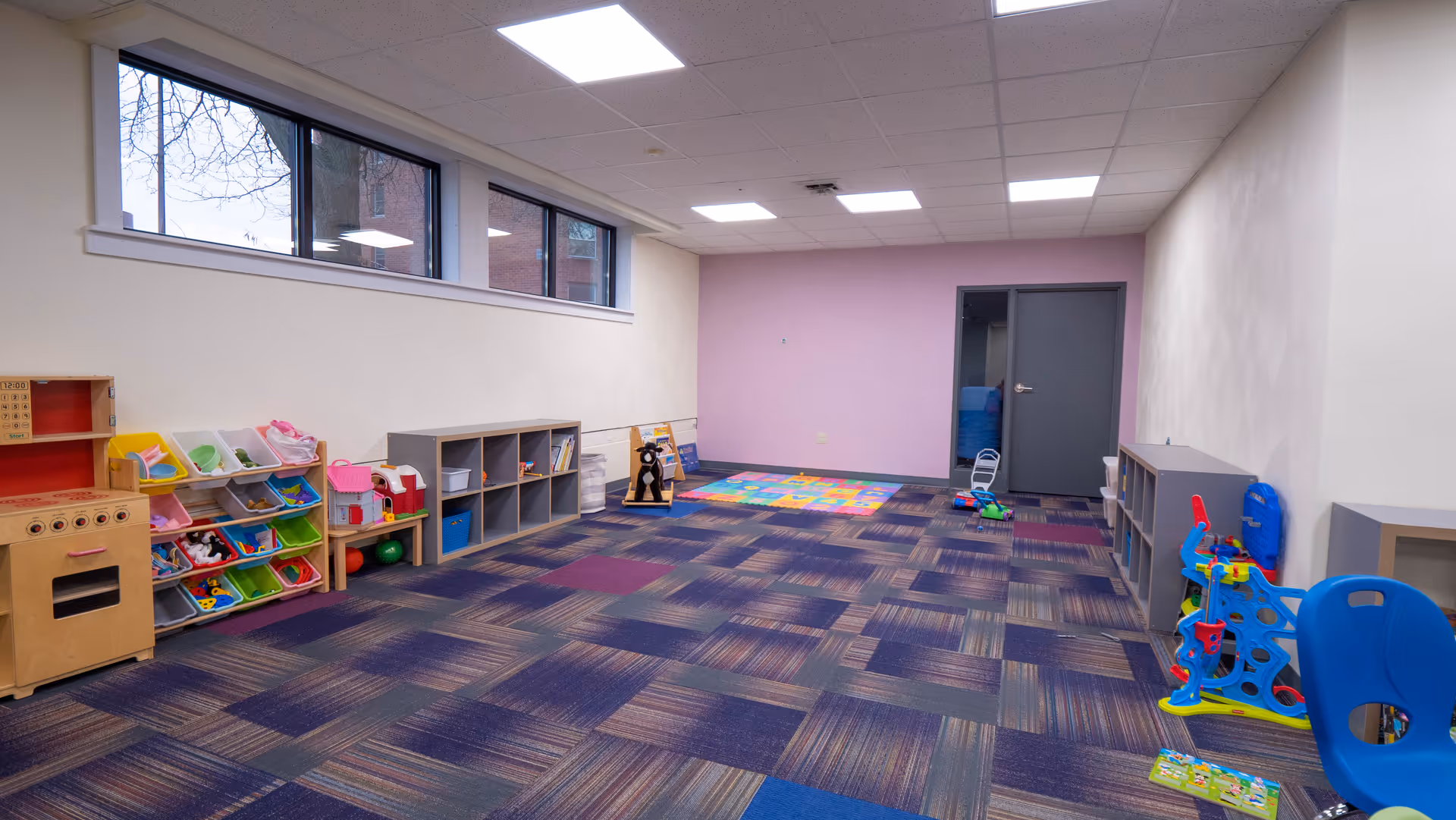 Bright indoor playroom with toy shelves, mats, and colorful carpet.