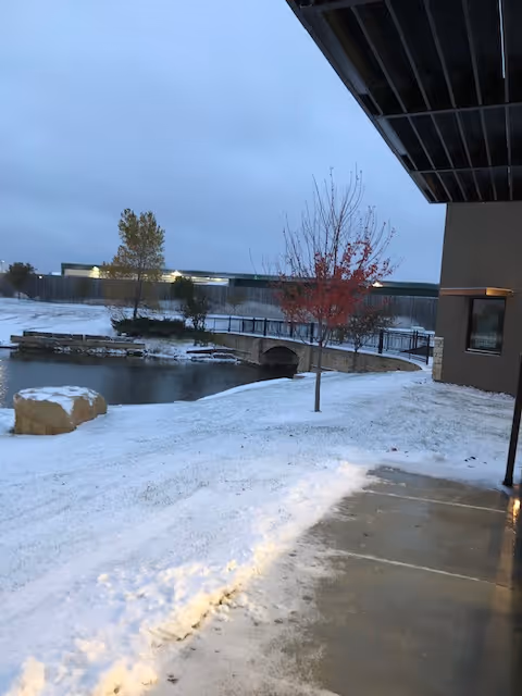 Snow covers a walkway beside a building, with a pond, bridge, and bare trees nearby.