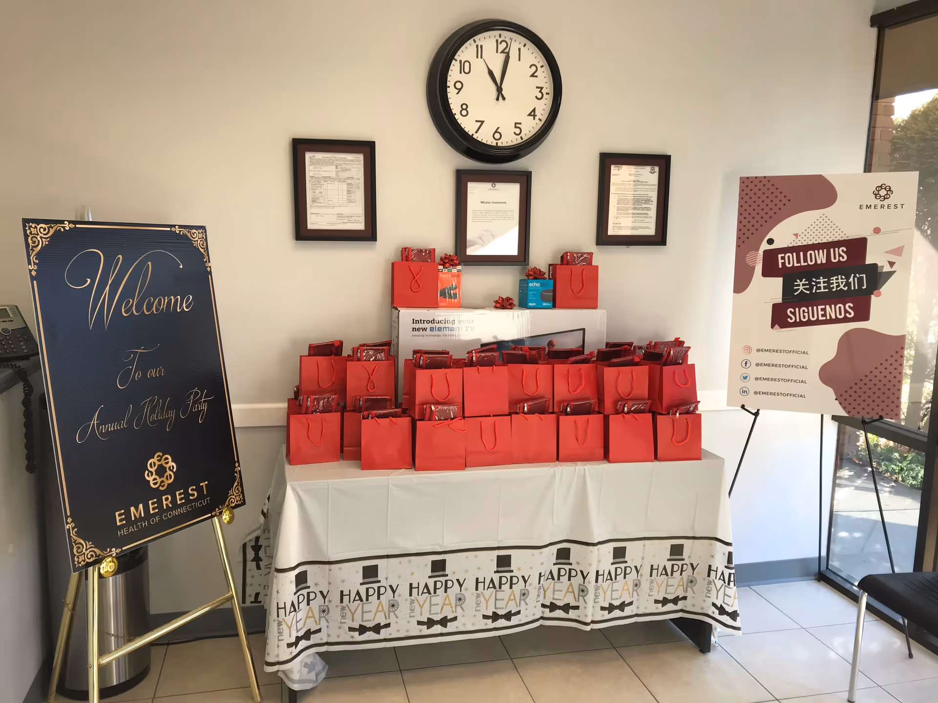 Indoor lobby table with red gift bags, clock on wall, welcome sign, and poster.