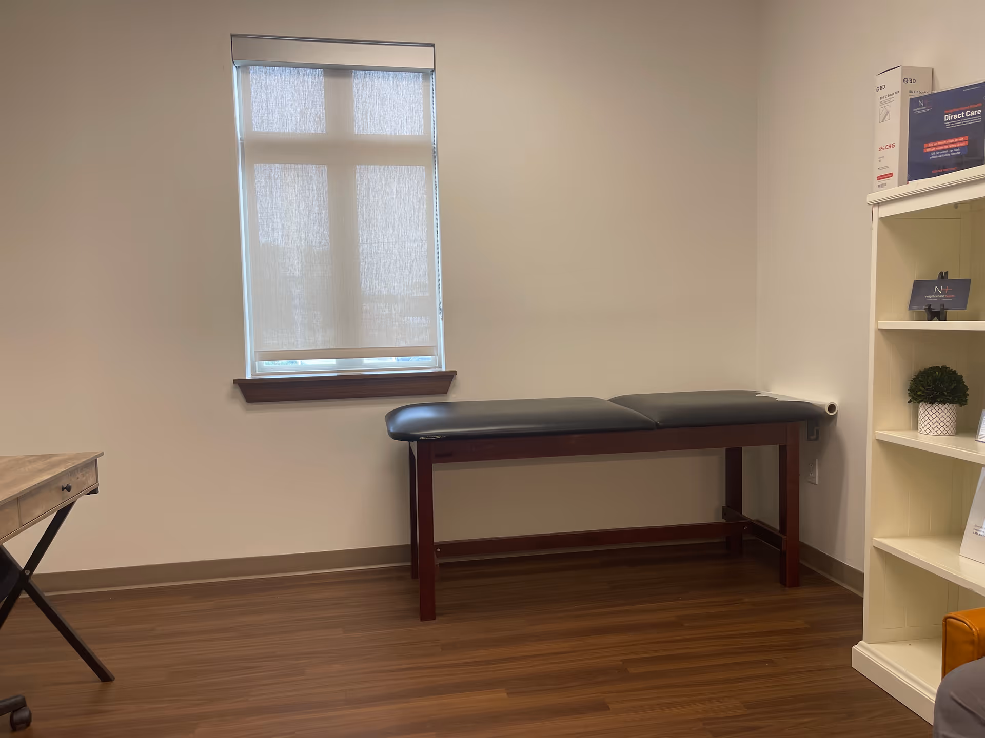A small clinical examination room with a padded table, window, and shelves.