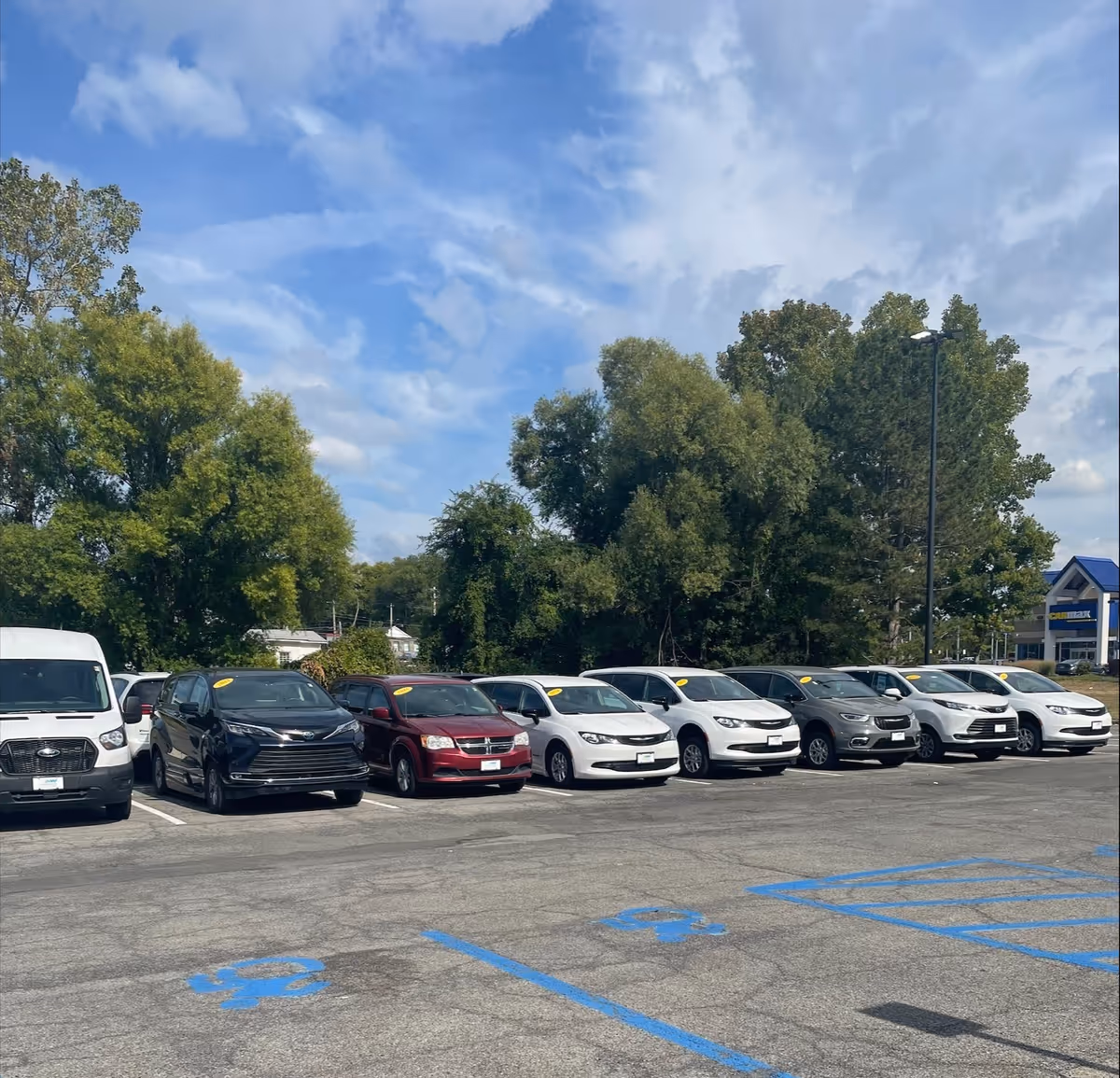 Row of cars parked in a lot beside trees, with a distant building in the background.