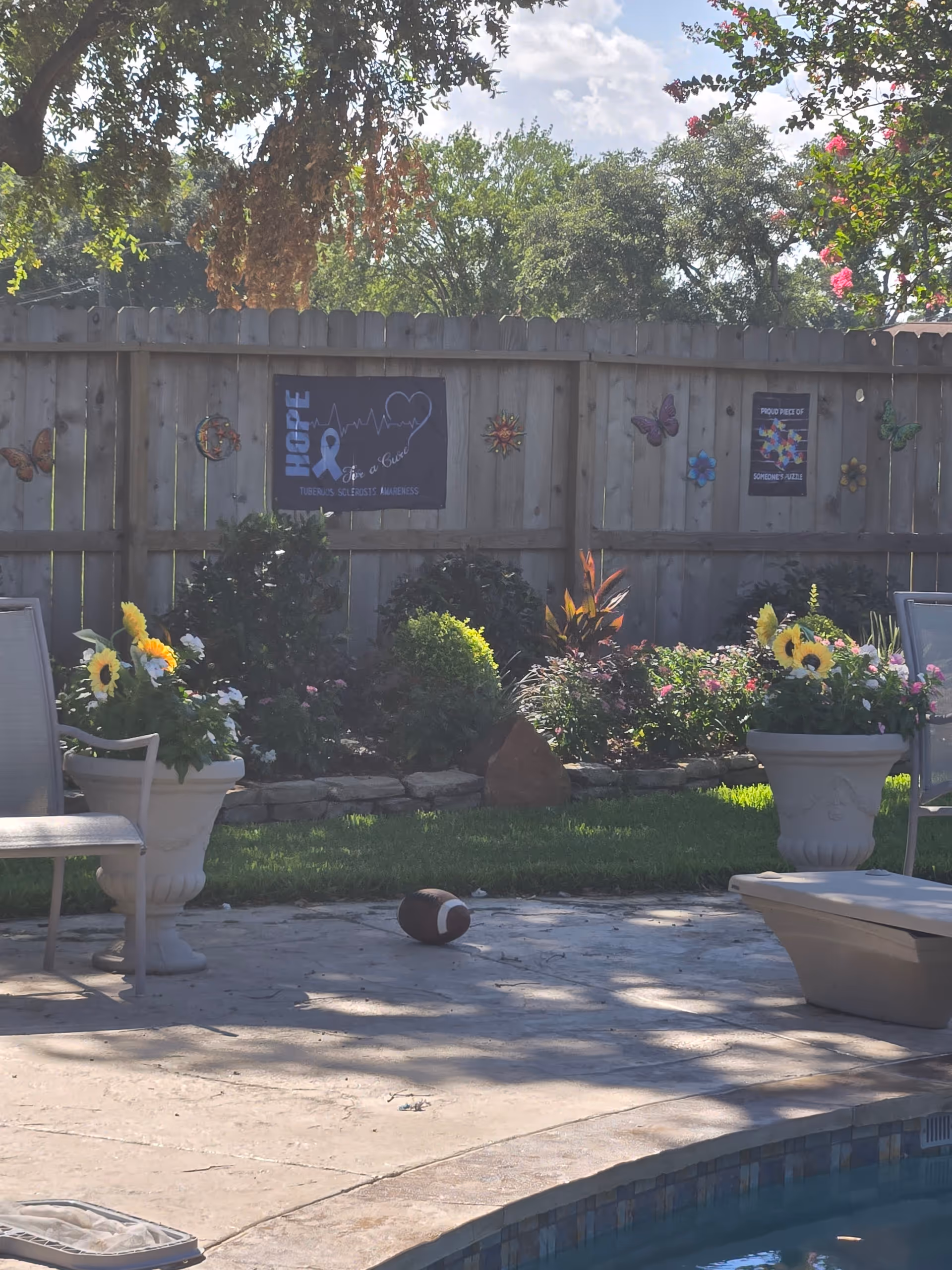 Sunlit outdoor courtyard with potted flowers, chairs, and a pool by a wooden fence
