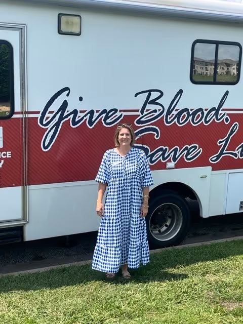 A woman in a blue and white check dress stands beside a red blood drive bus.