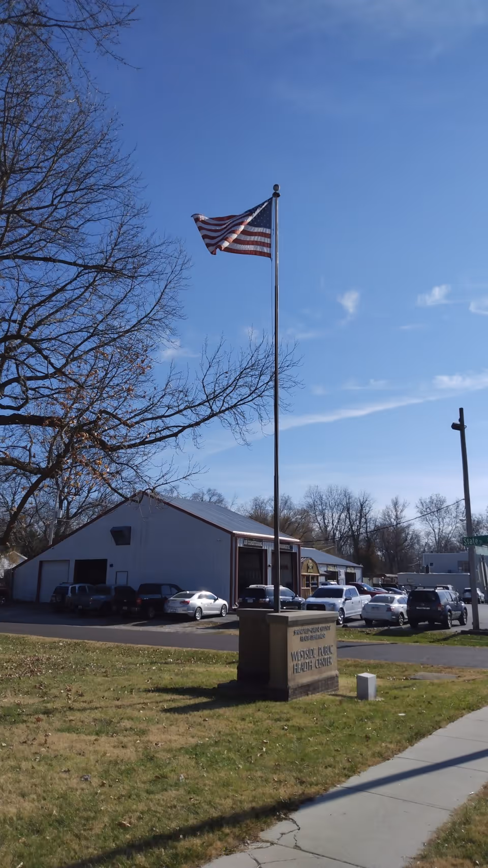 American flag on a tall pole in front of a public health center with parked cars.