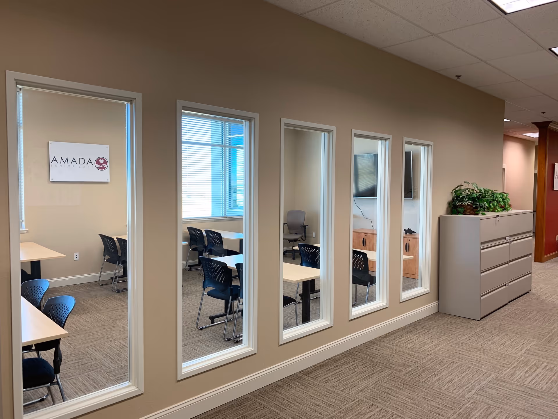 Hallway with windowed rooms showing desks, chairs, and an Amada Senior Care sign.