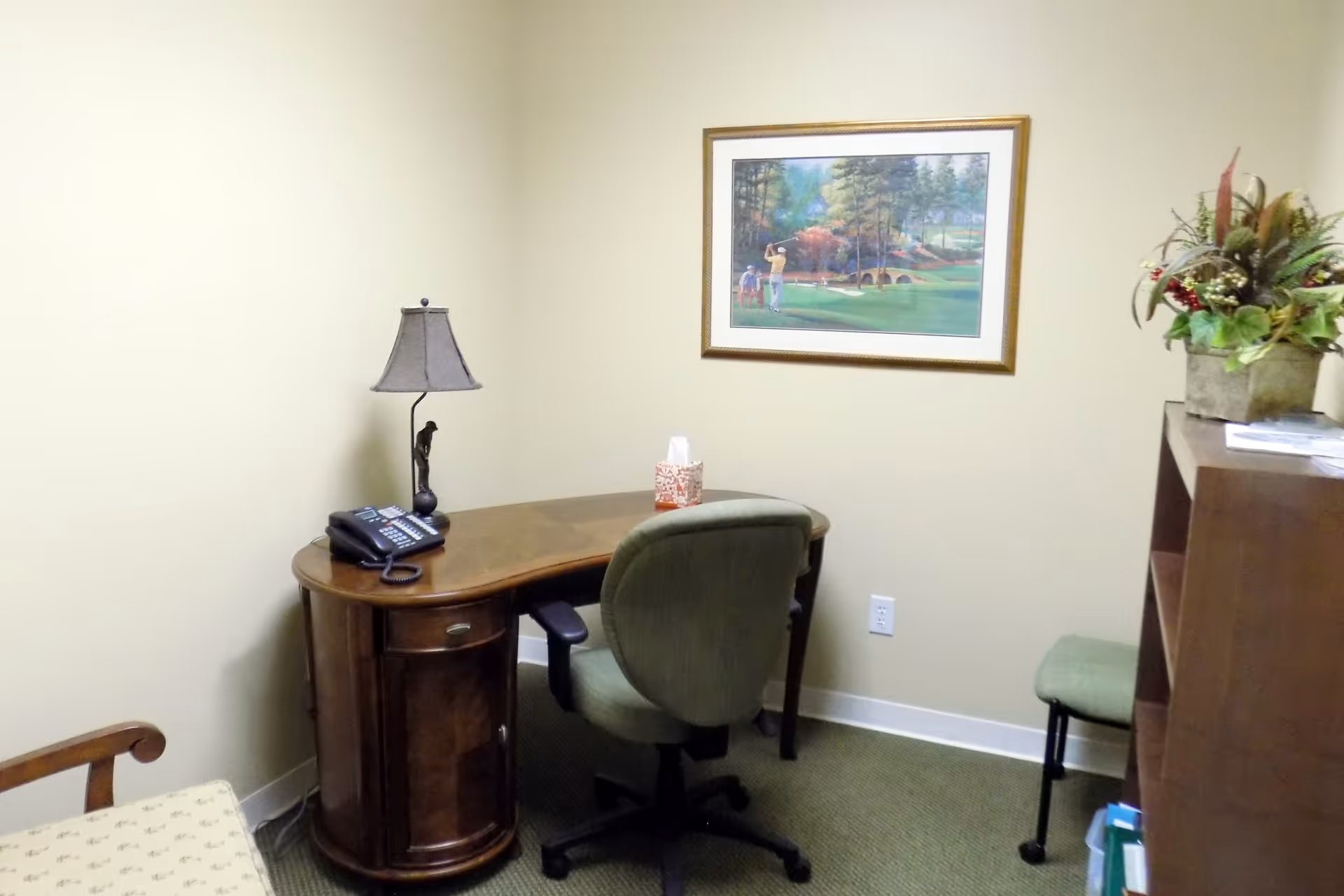 Small office corner with wooden desk, chair, lamp, artwork, and a shelf.
