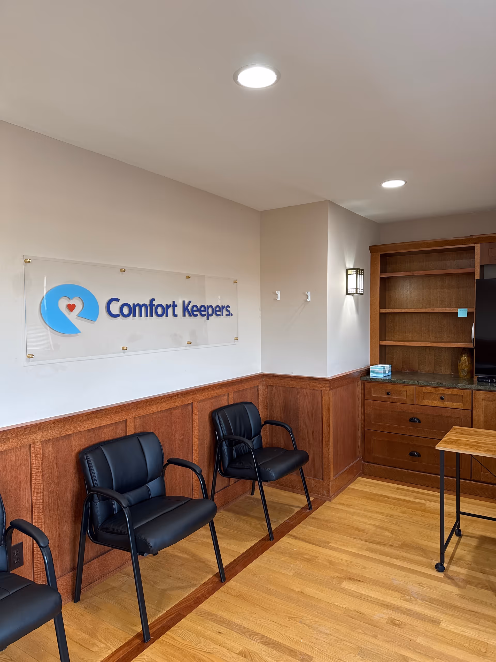 A reception lobby with Comfort Keepers sign, wood wainscoting, and black chairs.
