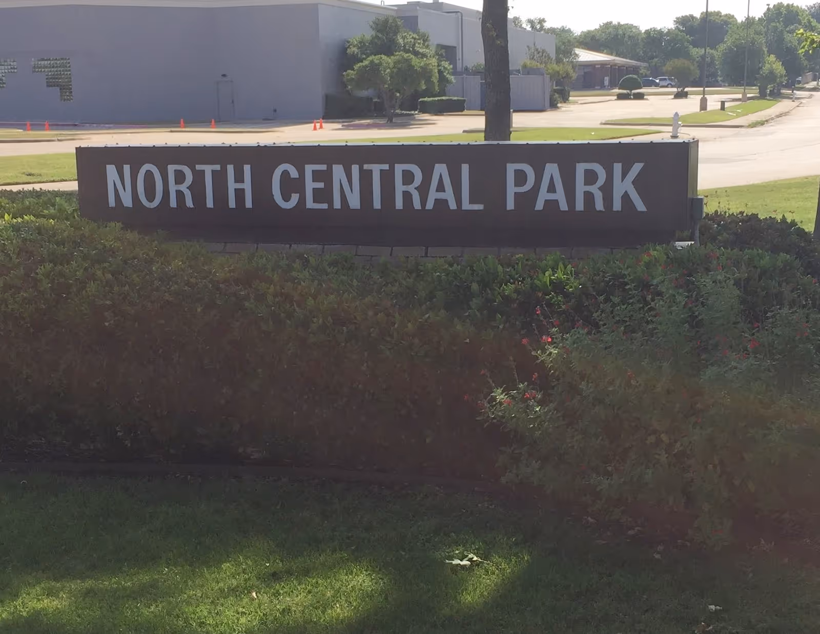 Large outdoor park sign reading North Central Park with shrubs and a road in the background.