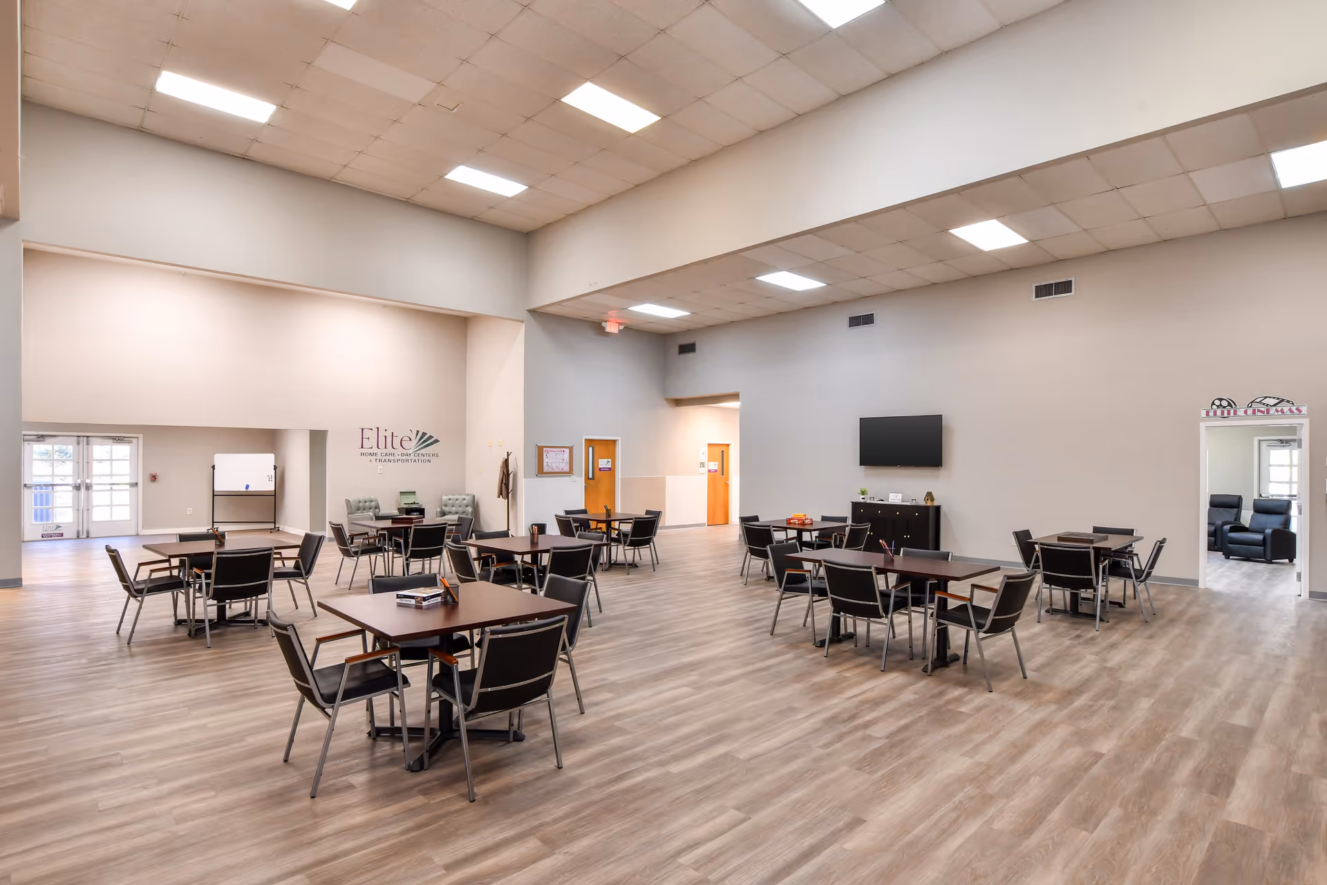 Spacious indoor senior living dining hall with multiple tables, chairs, and a wall-mounted TV.