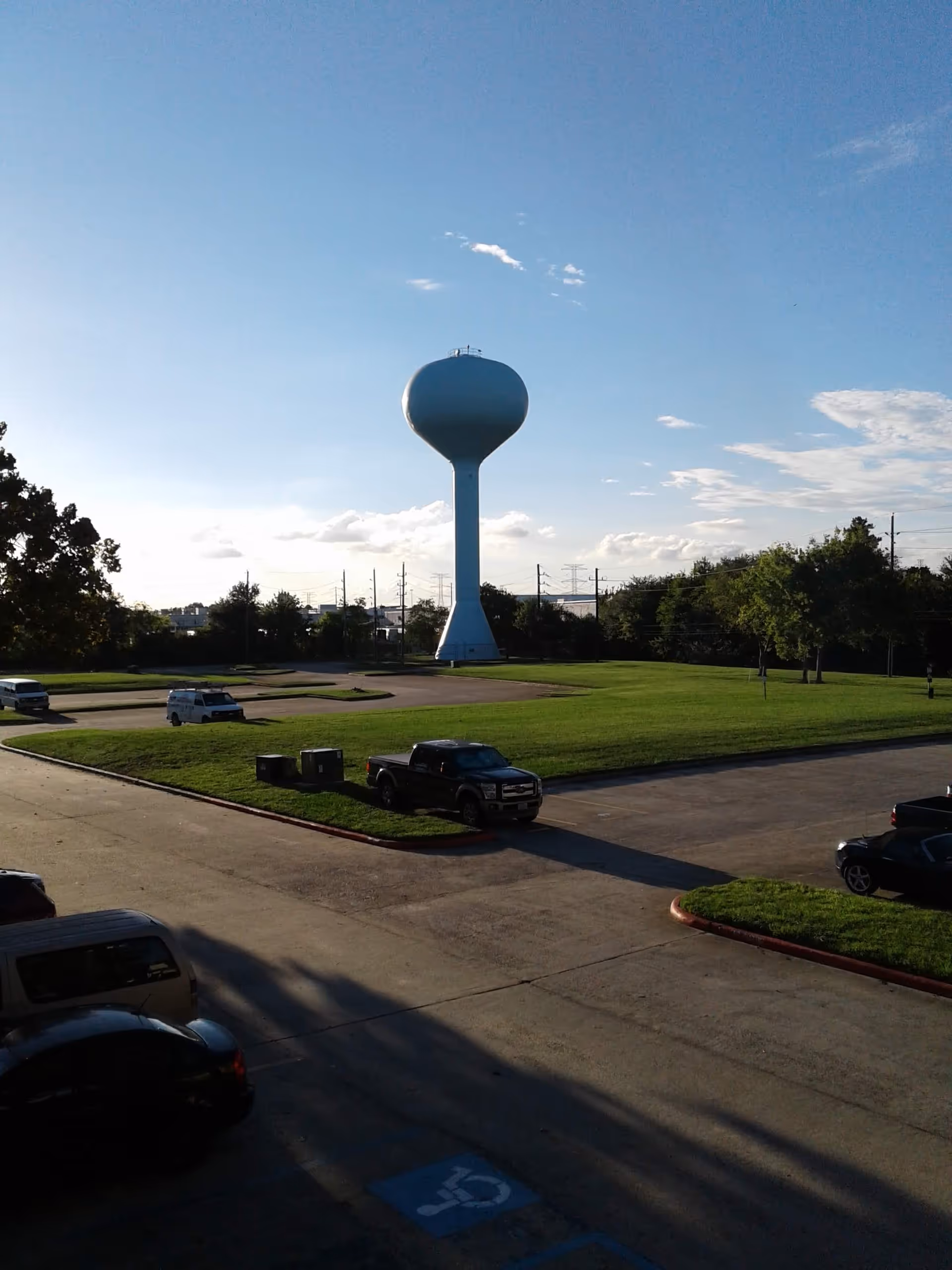 Wide outdoor scene featuring a tall white water tower over green lawn and parked cars.