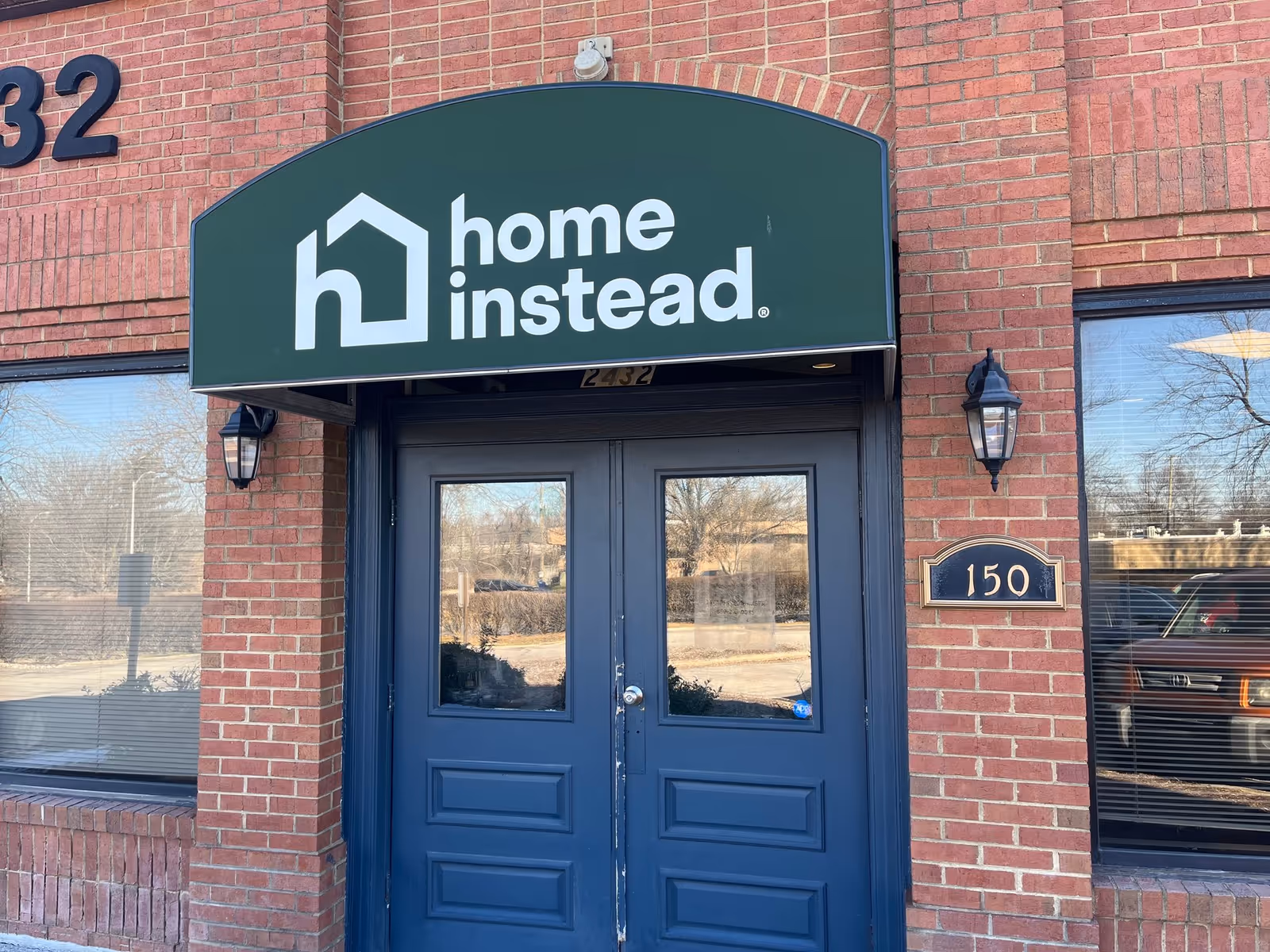 Brick building entrance with blue double doors and a green Home Instead sign.
