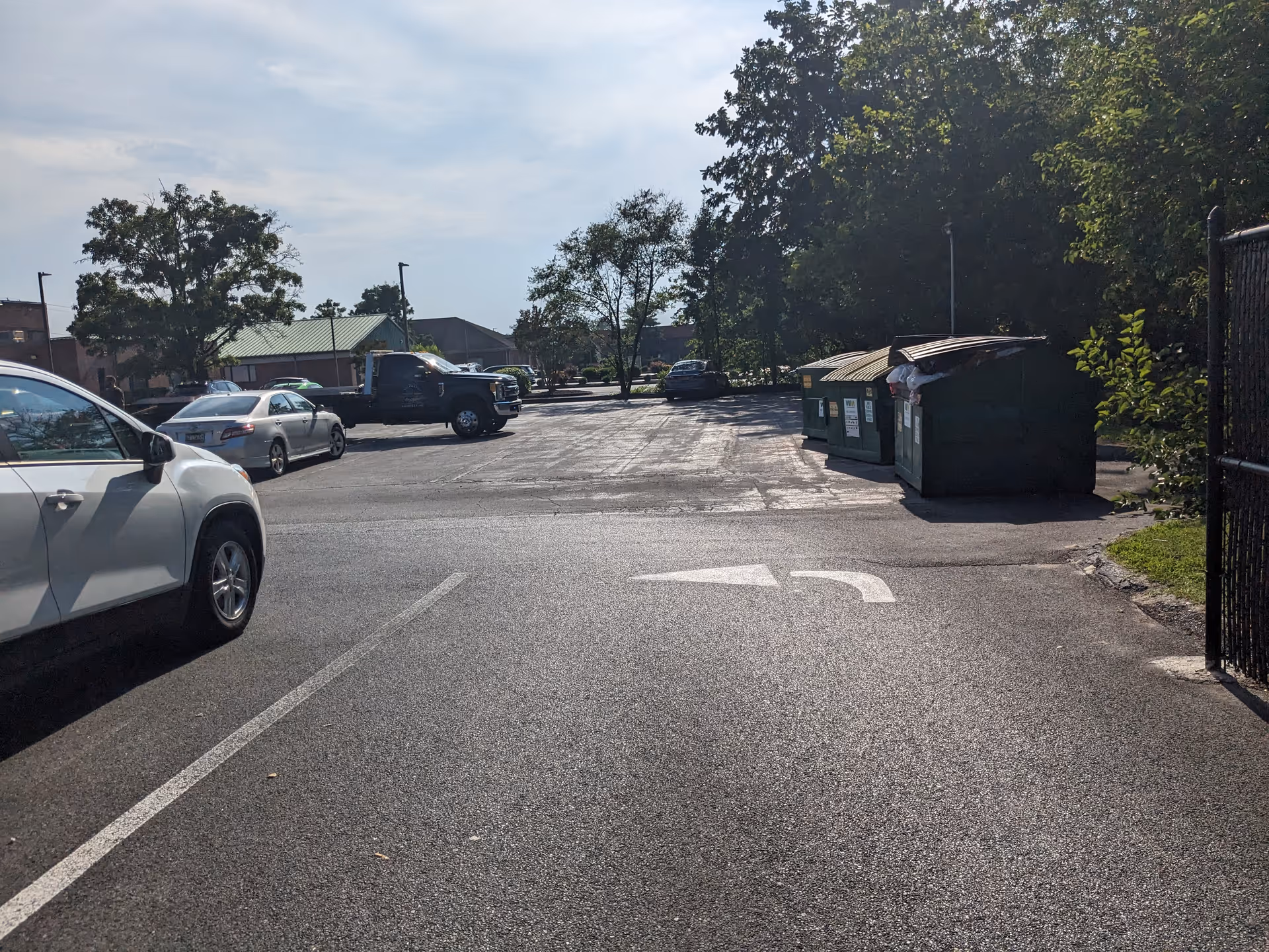 An exterior parking lot with parked cars, dumpsters, and trees along a gated area.