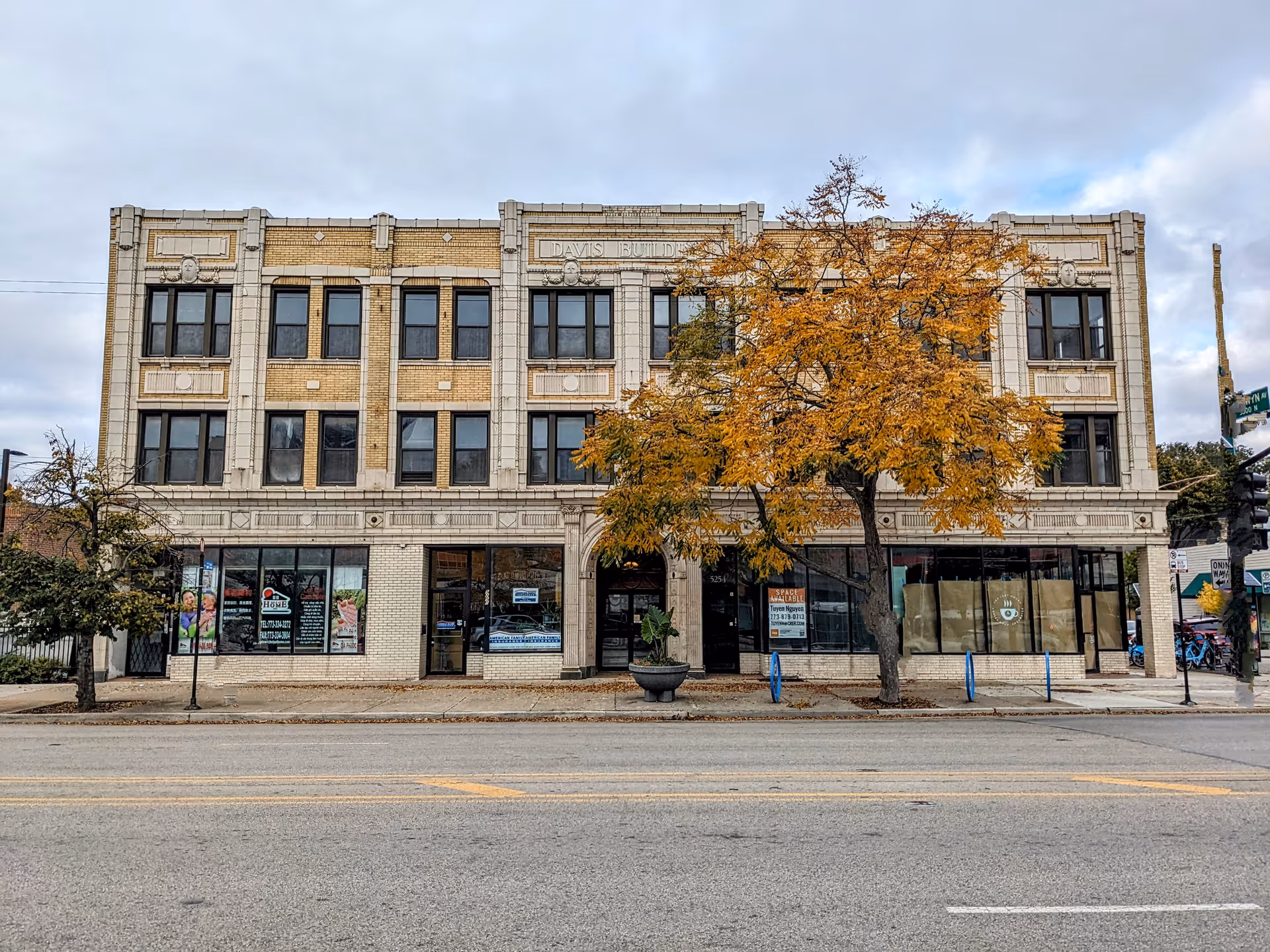 Four-story brick building with large storefront windows and a tree-lined sidewalk.