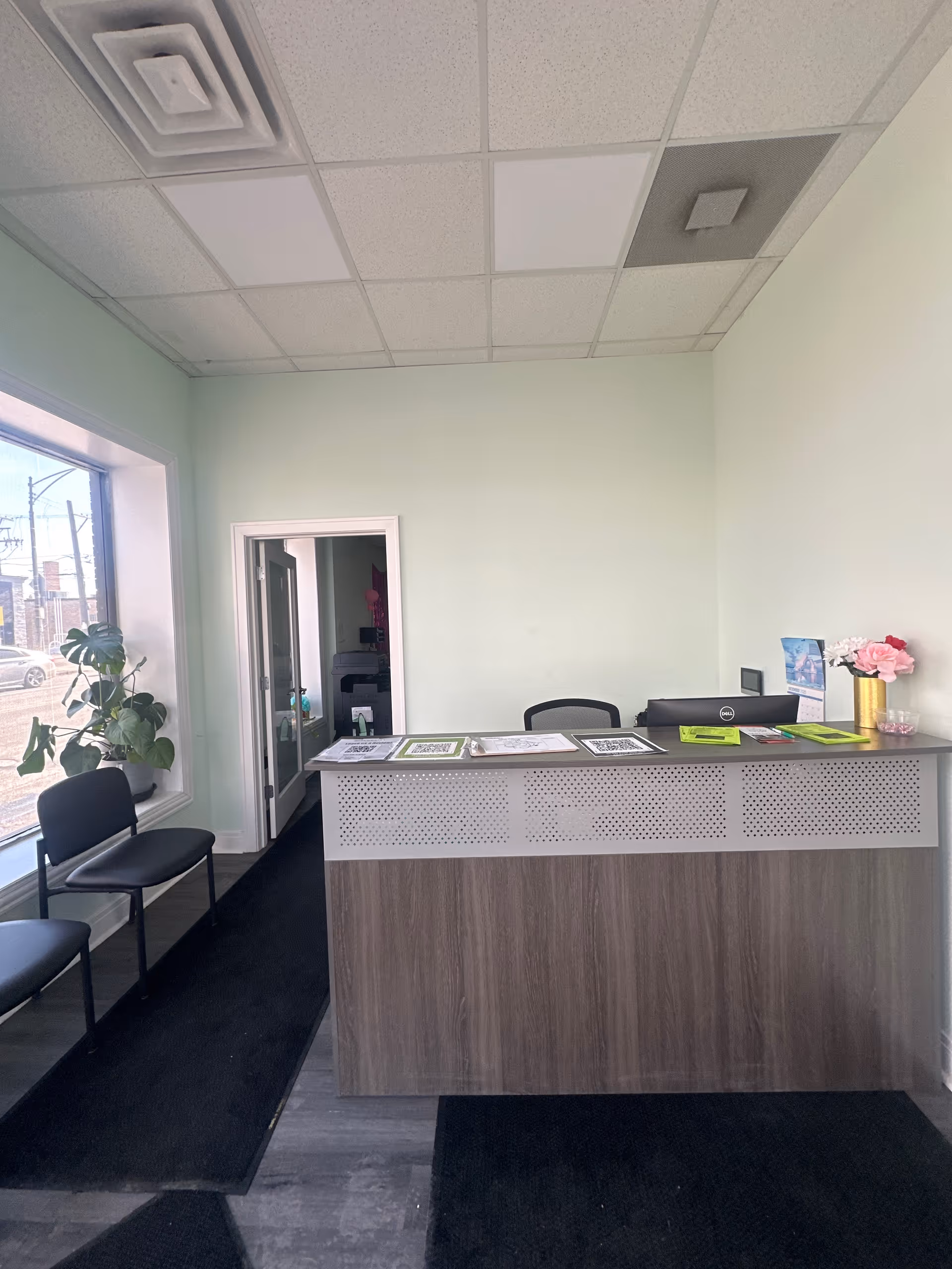 Reception desk in a bright lobby with chairs, a window view, and a plant.