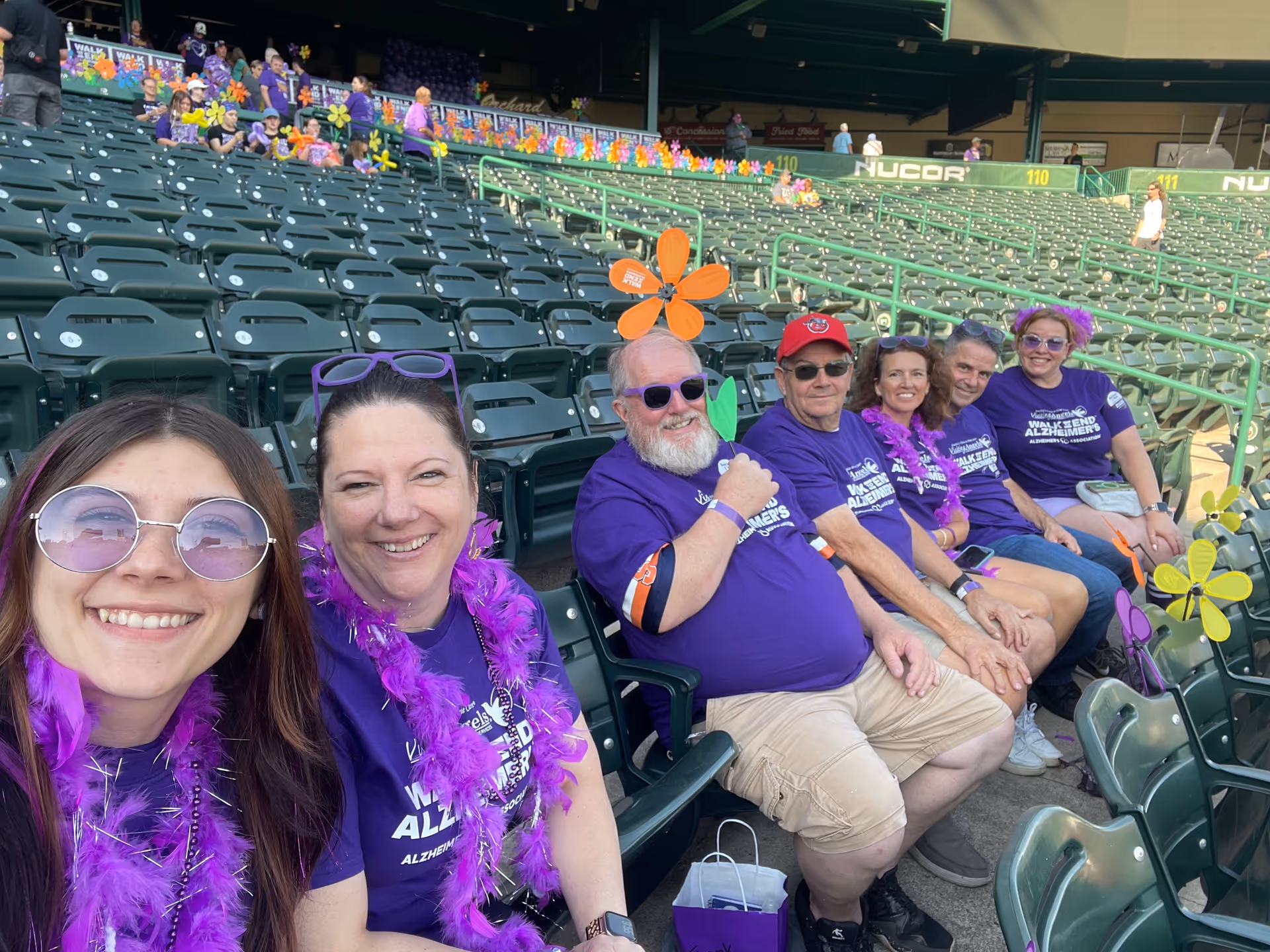Group of seniors and caregivers in purple shirts sit together in stadium seats.