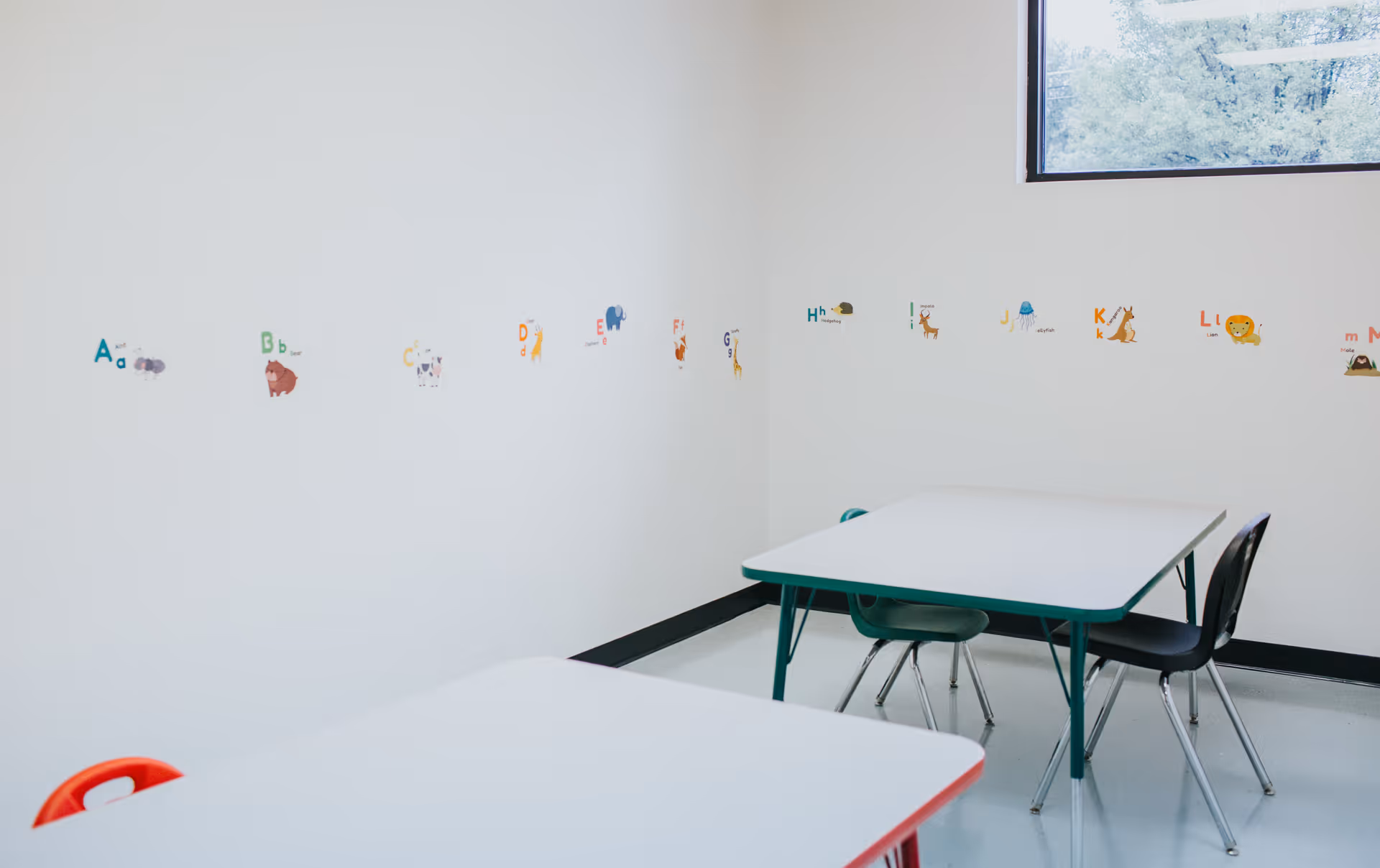 An empty classroom-style room with small tables, chairs, and alphabet decals along white walls.