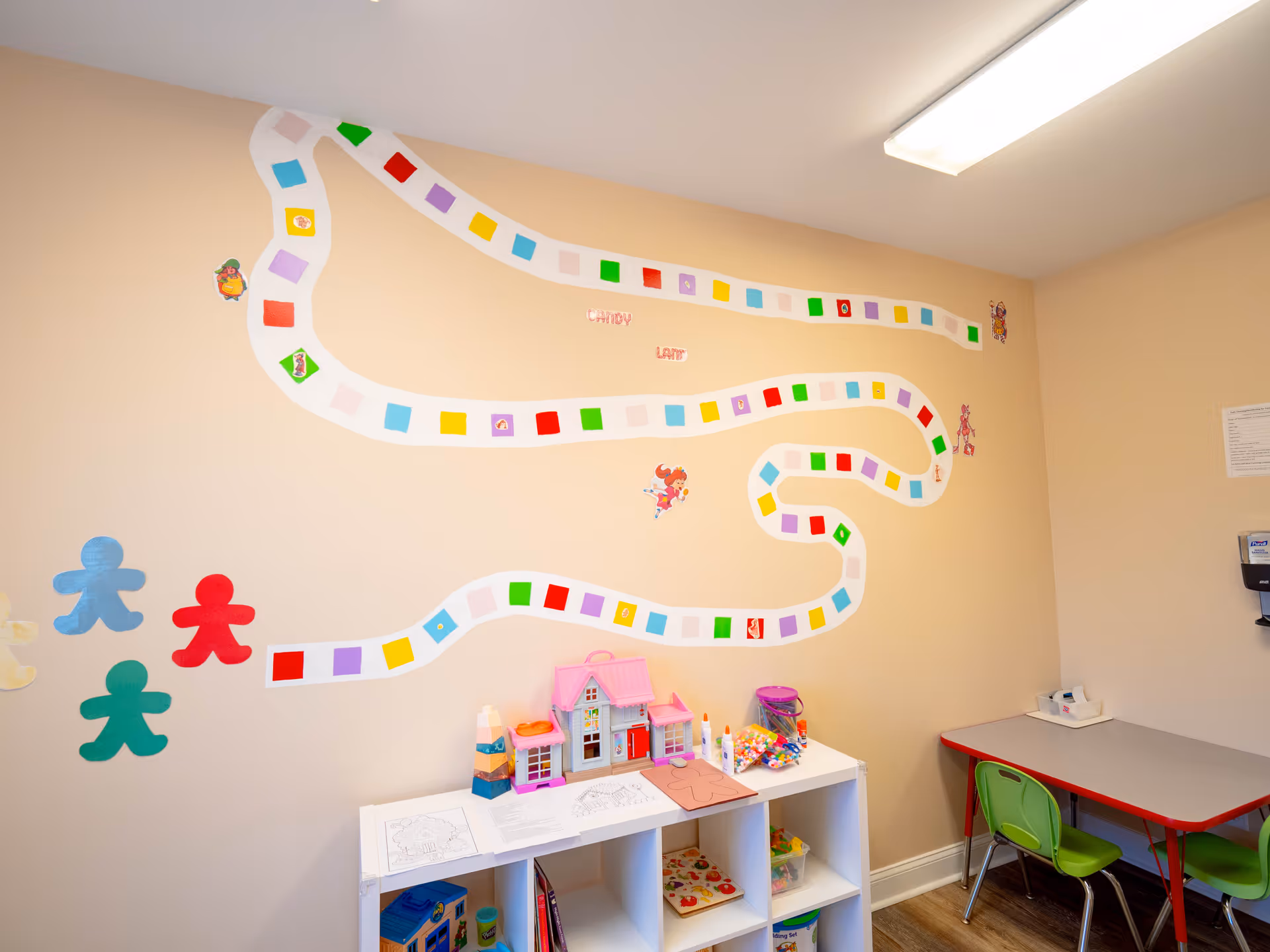 Playroom with a winding color-path mural, toy house, shelf of toys, and small table with chairs.
