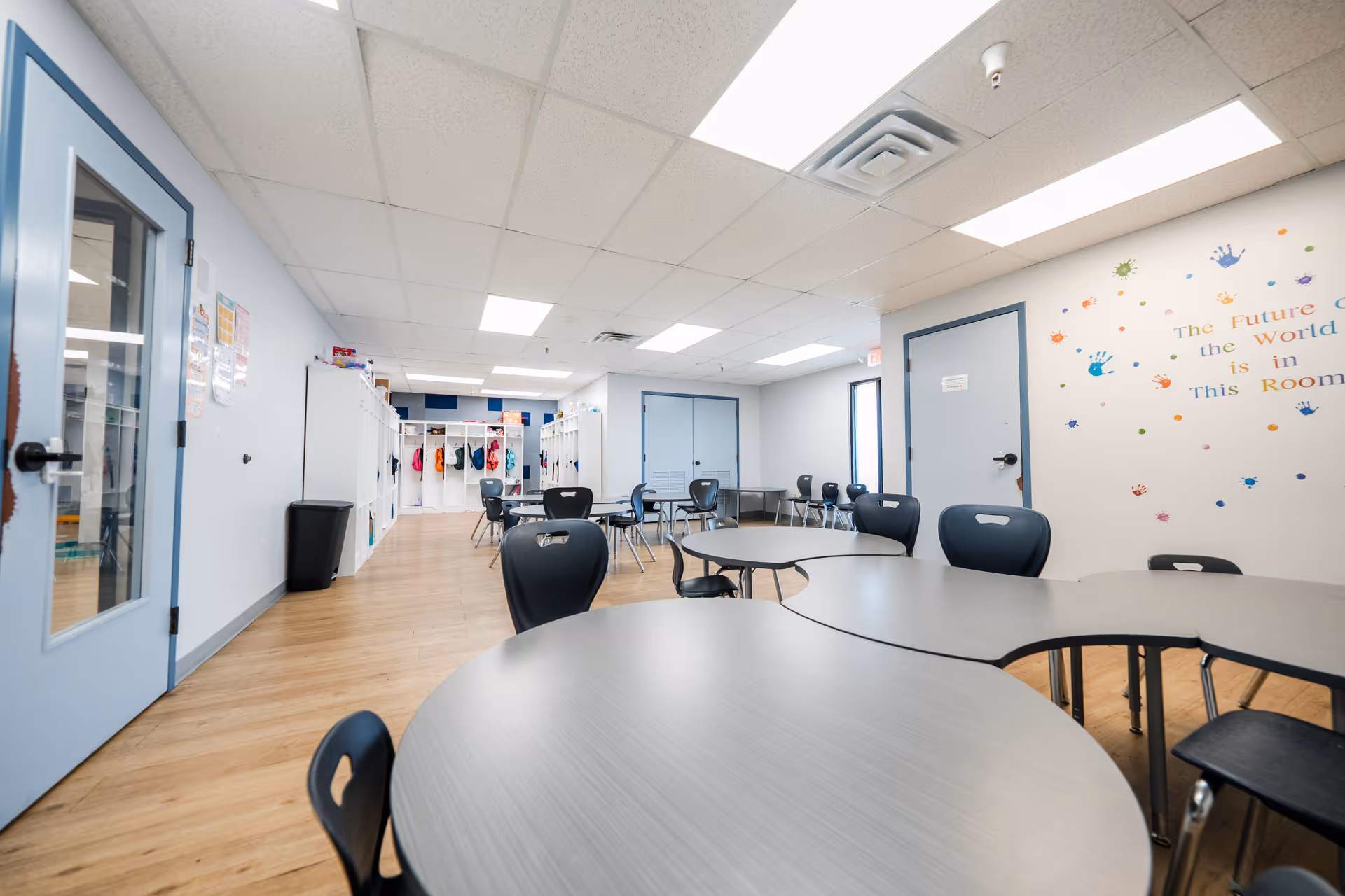 Bright interior activity room with round tables, chairs, and lockers along the back wall.