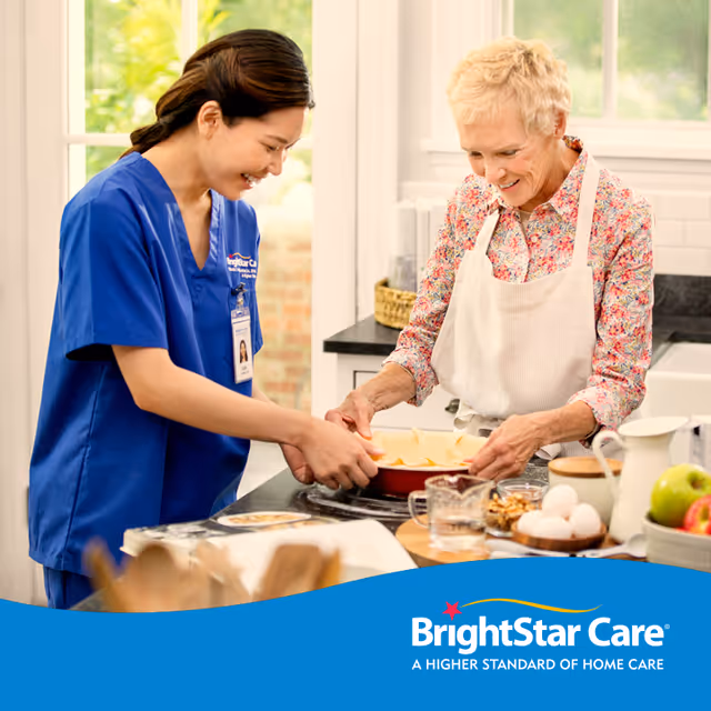 A caregiver in blue scrubs helps an elderly woman cook in a bright kitchen.