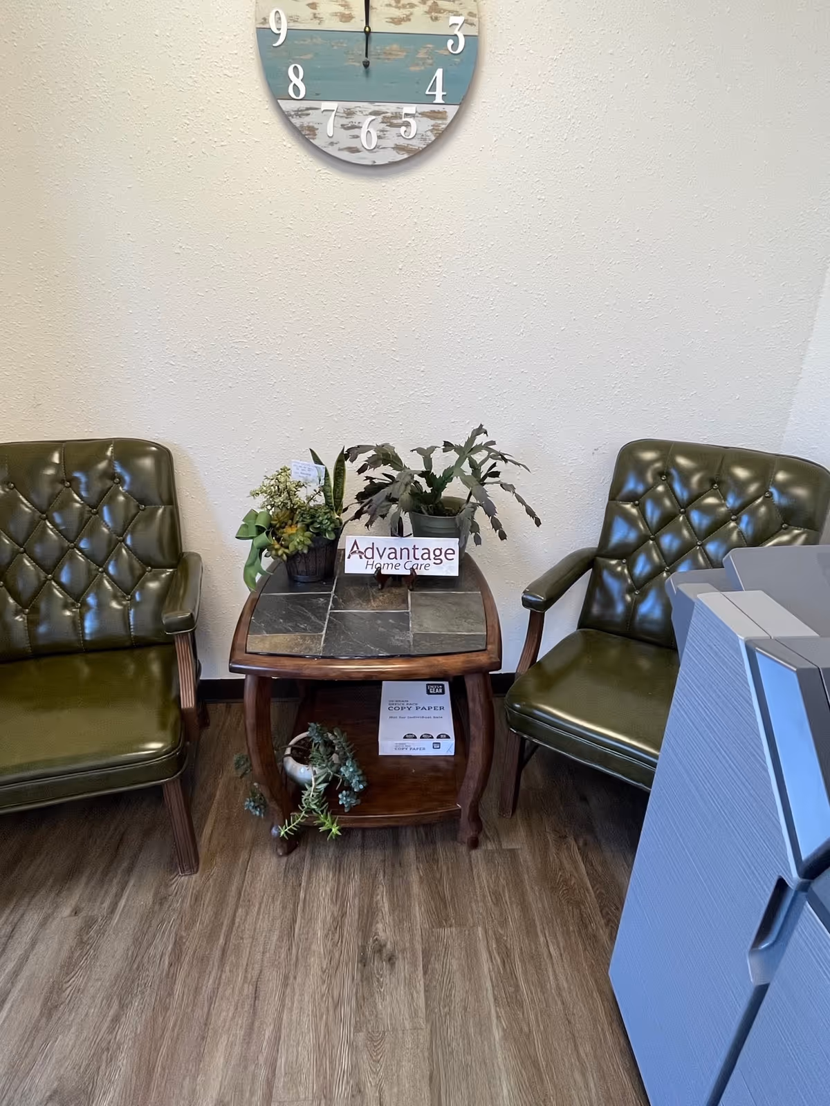 Lobby seating area with two green tufted chairs, table, plants, clock, and Advantage Home Care sign.