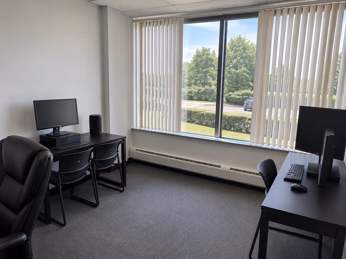 Office workspace with two desks, computers, and vertical blinds by a large window.