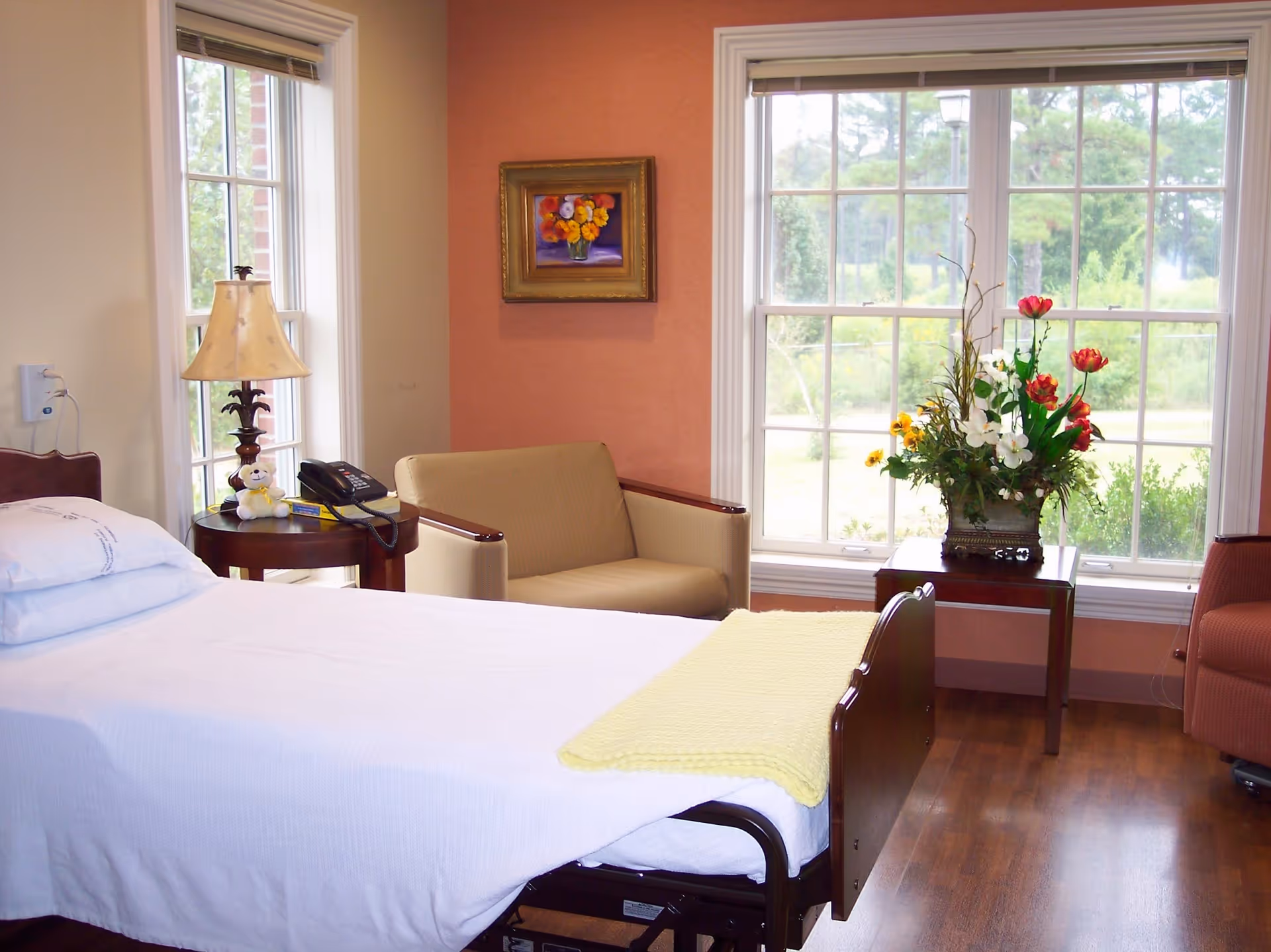 A hospital-style bed in a sunny patient room with chairs, table, and vase of flowers.