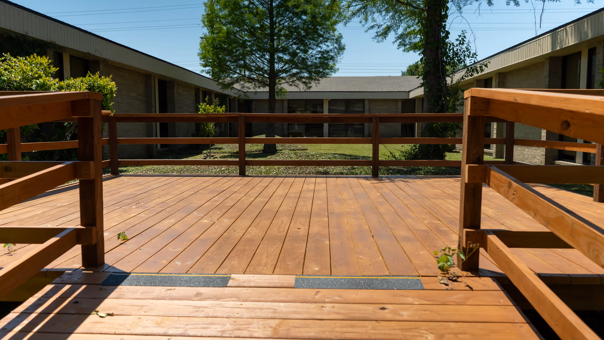 Wooden outdoor deck with railings overlooks brick buildings and green trees.
