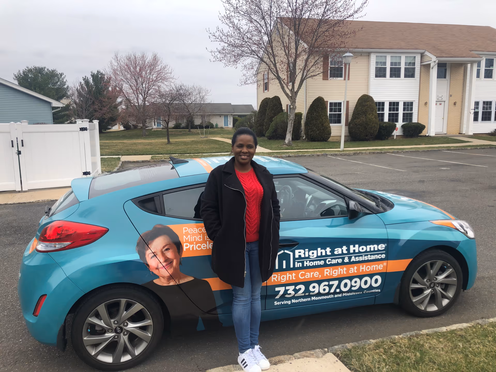 A smiling woman stands beside a teal Right at Home service vehicle in a parking lot.