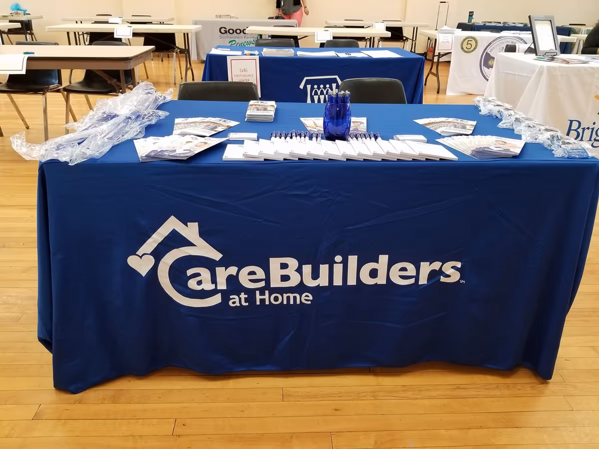 Blue booth table with CareBuilders at Home branding and promotional materials in a conference hall.
