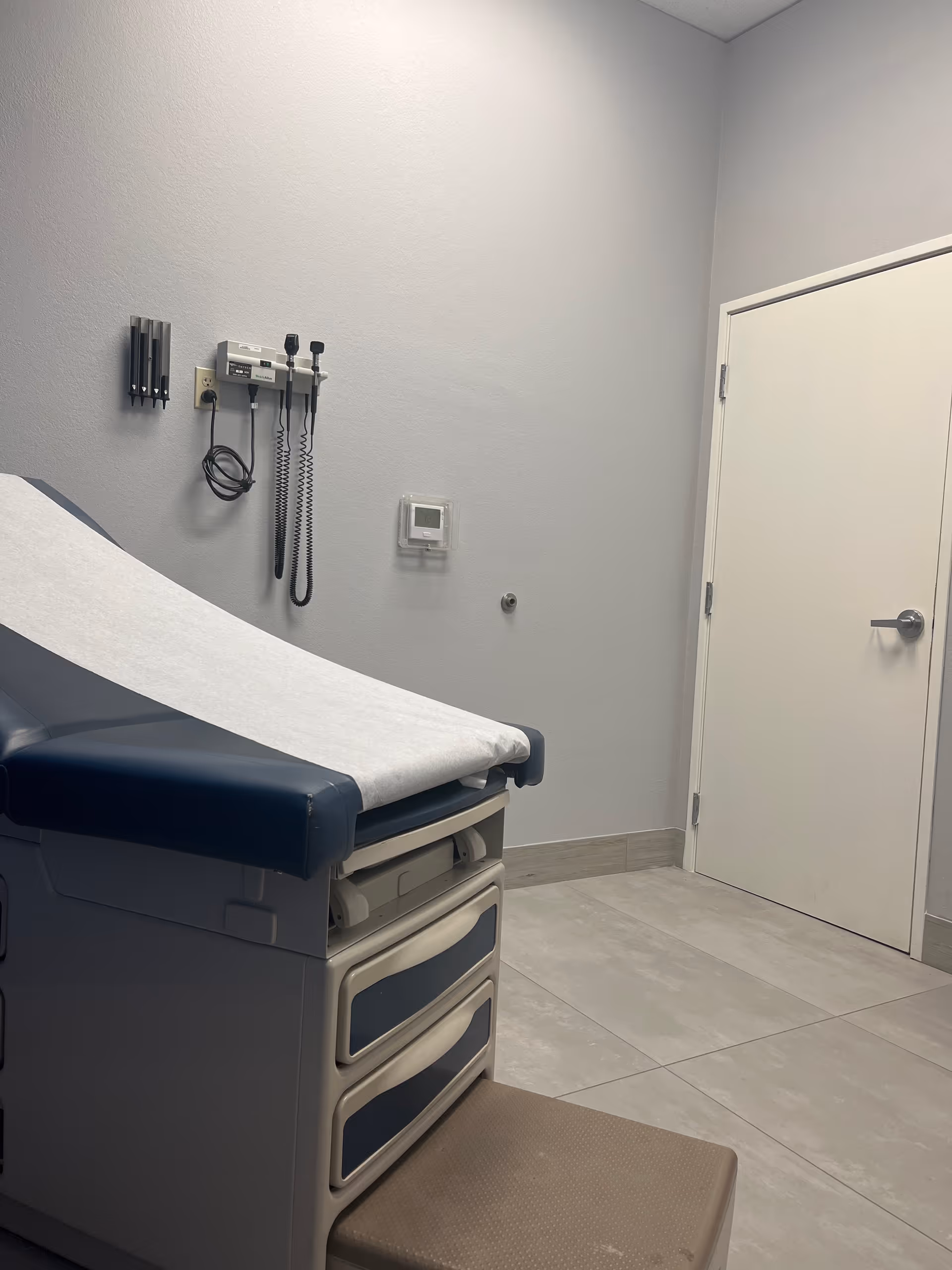 Medical examination room with exam chair, wall-mounted instruments, and closed door.