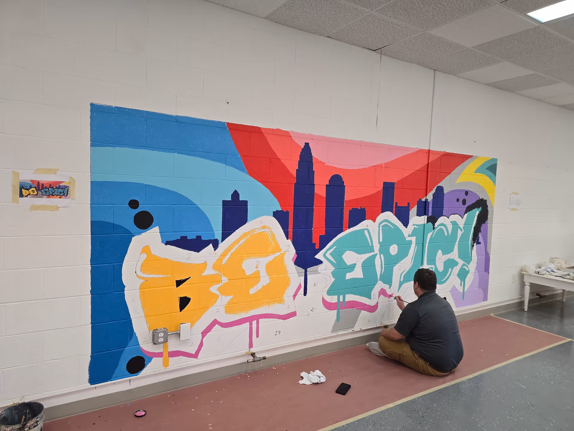 Person sits on the floor painting a vibrant mural on a white brick wall indoors.