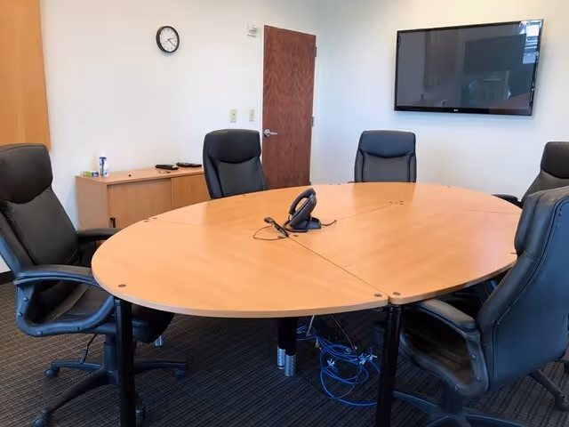 Round wooden conference table with black leather chairs in a meeting room with TV.