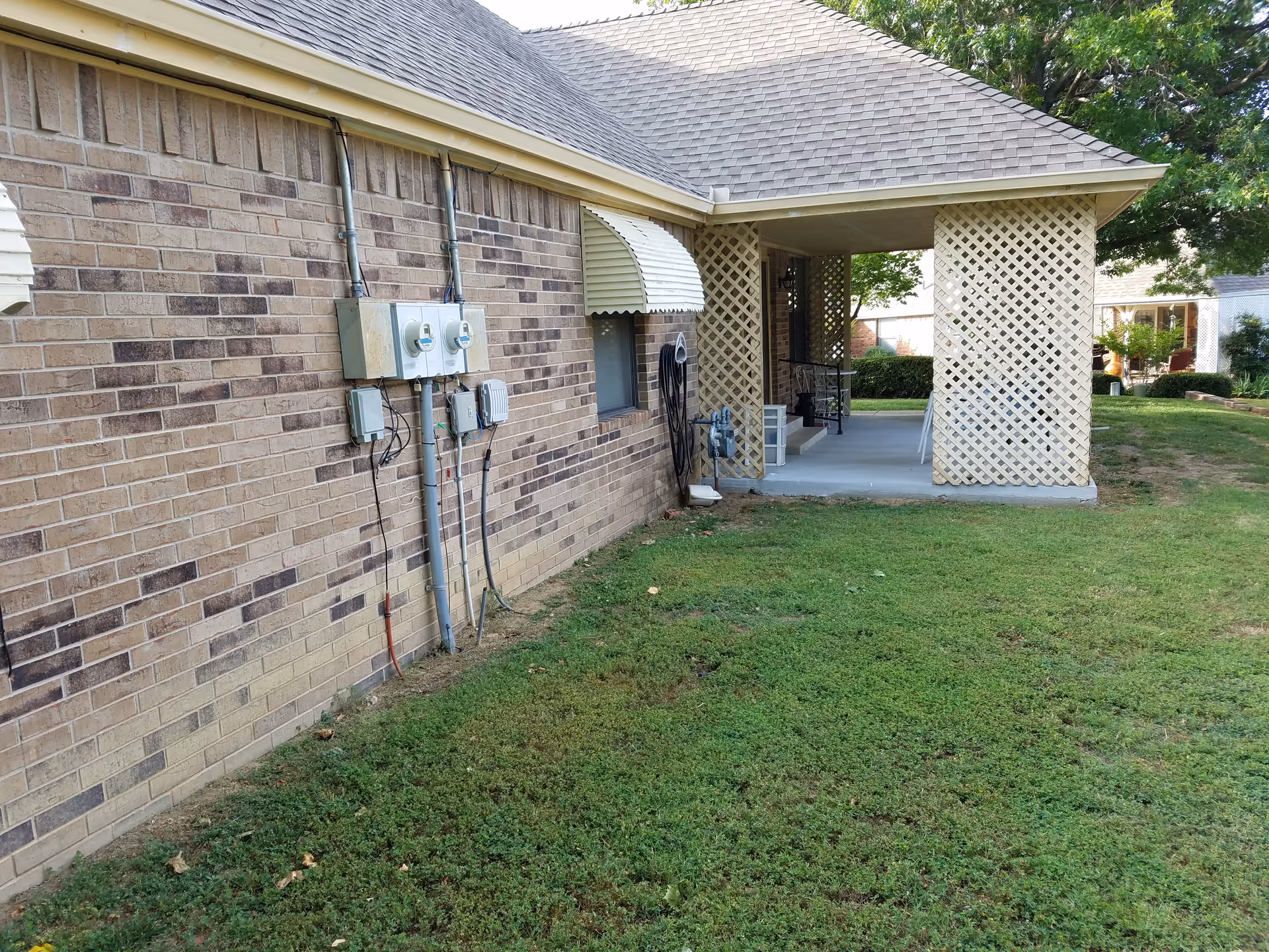 Brick exterior with a covered lattice patio, utility meters, and a grassy yard.