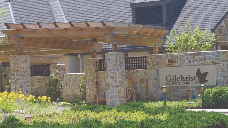 Stone entrance with wooden pergola and Gilchrist Hospice Care sign.