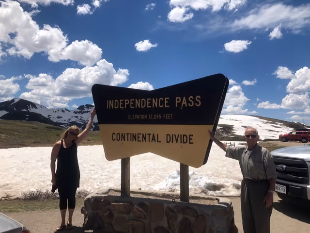 Two people stand beside the Independence Pass sign amid snow and mountains.