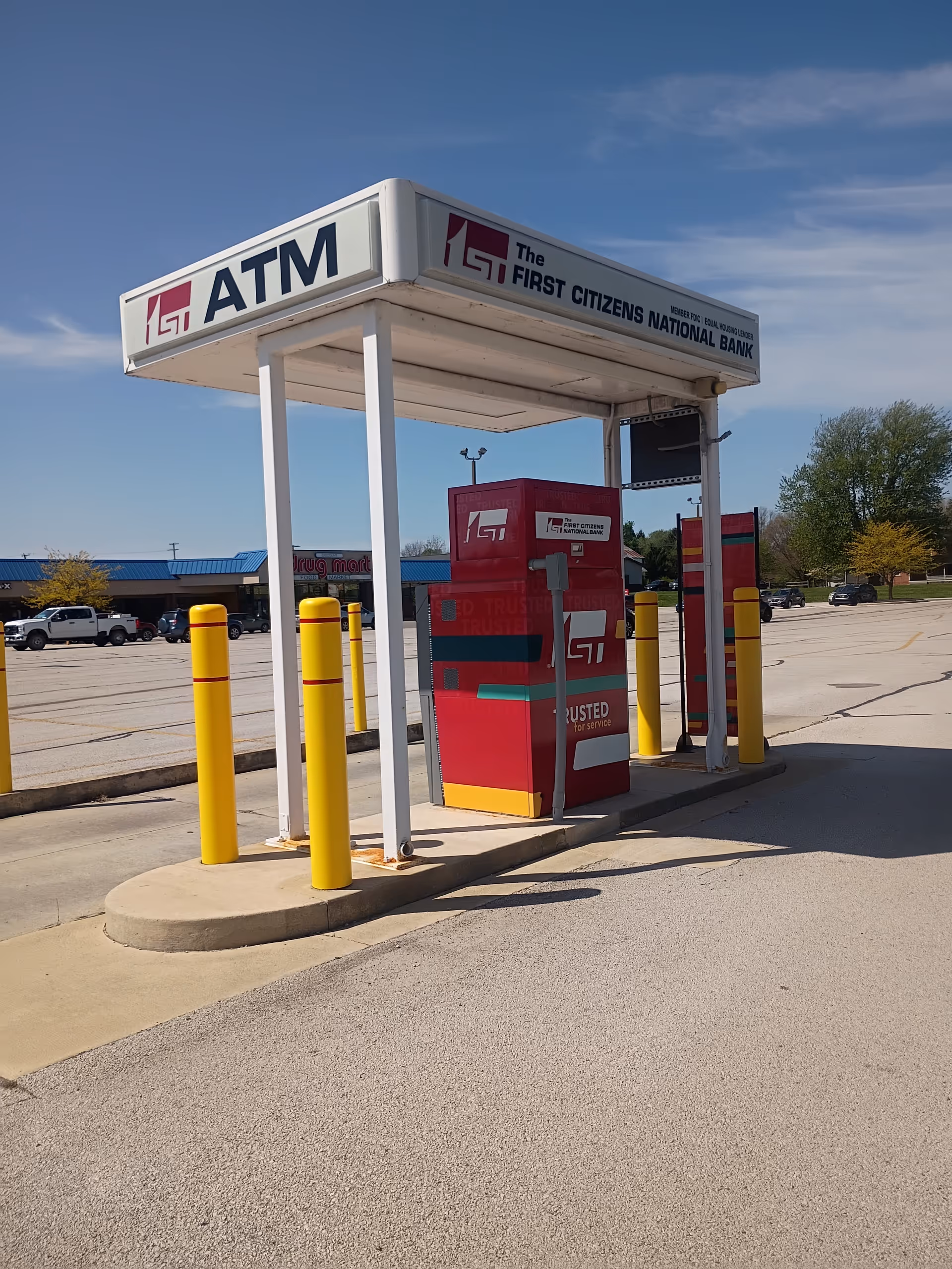 An outdoor ATM kiosk under a white canopy with a red machine and yellow bollards.