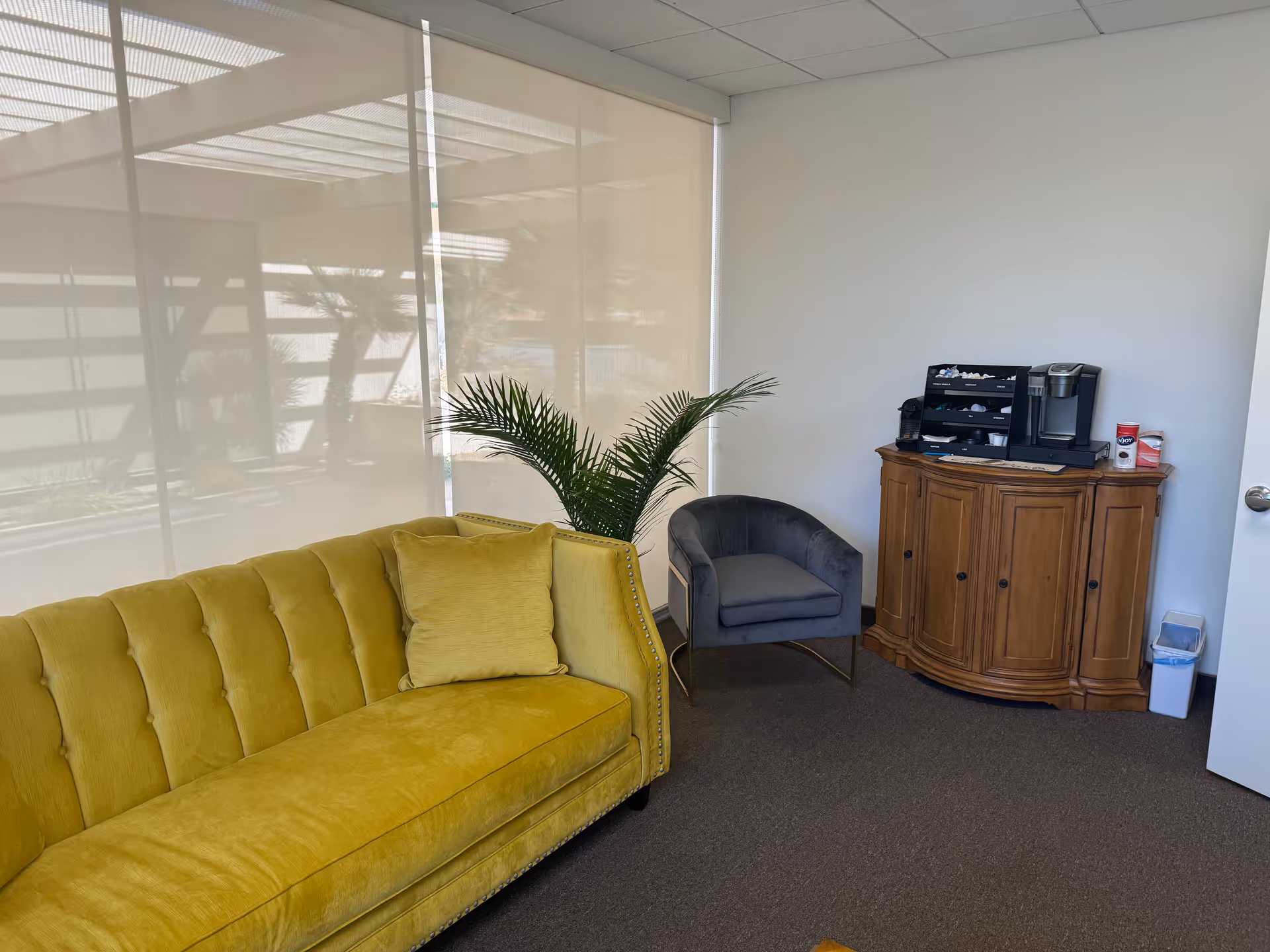 Bright interior lounge with yellow sofa, gray chair, plant, and wooden coffee station.