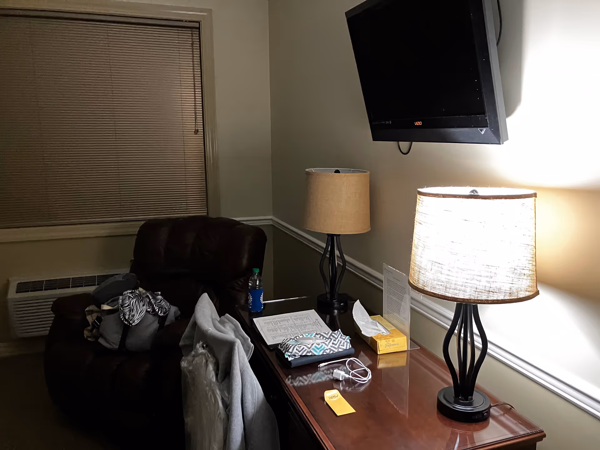 A dim living room with a wall-mounted TV, two lamps, and clutter on a wooden table.