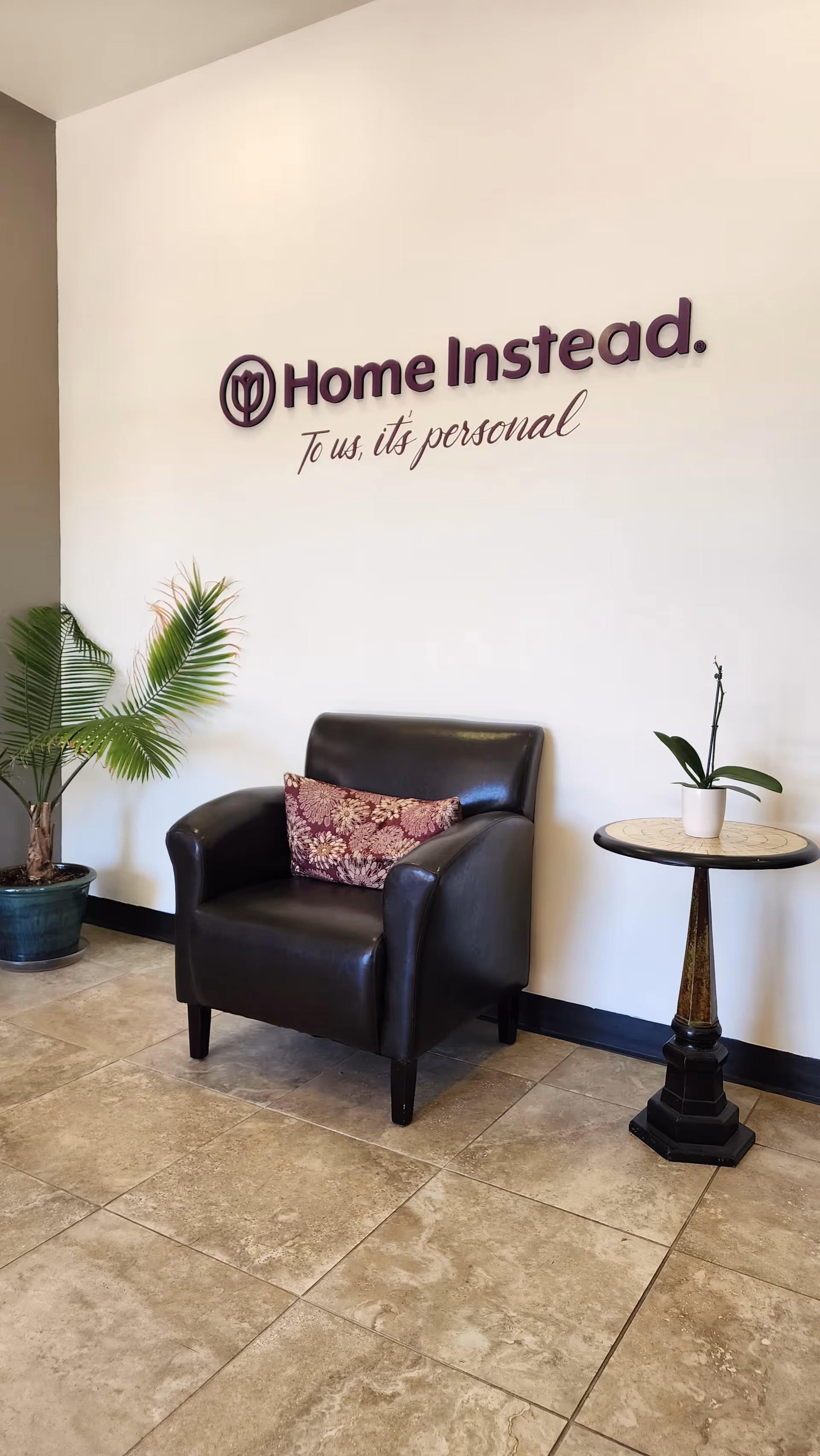 Interior lobby with a black chair, potted plants, and a Home Instead sign on the wall.