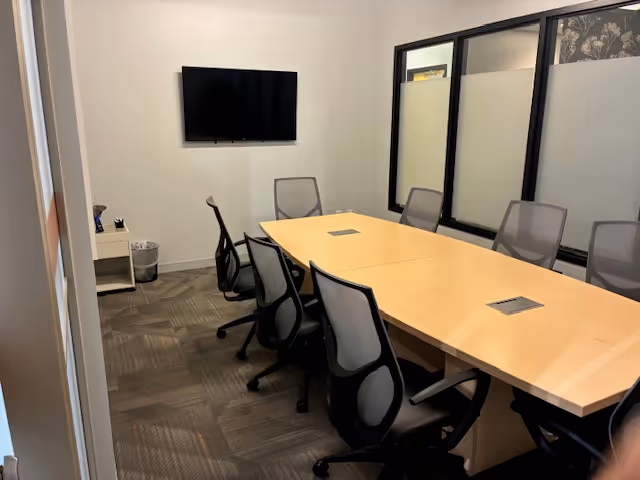 Conference room with long light-wood table, gray chairs, wall-mounted TV, and glass partitions.