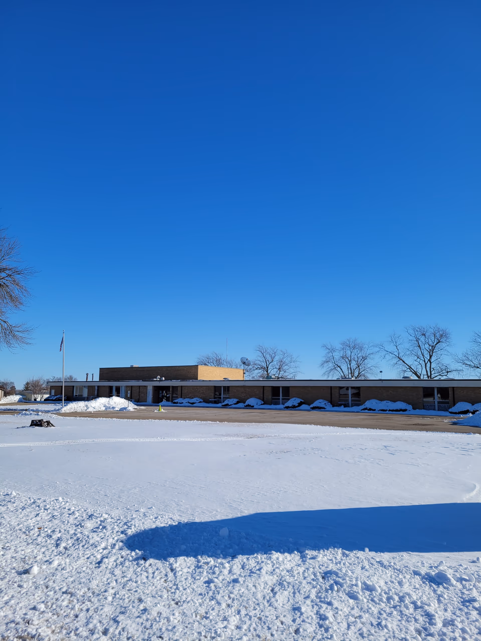 Snow-covered ground in front of a low, single-story Help at Home facility under clear blue sky.