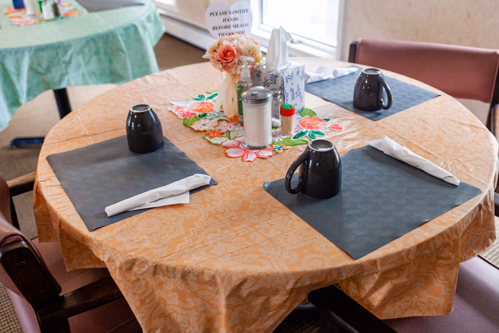 Round table with peach tablecloth, napkins, mugs, condiments, and flowers in a senior dining room.