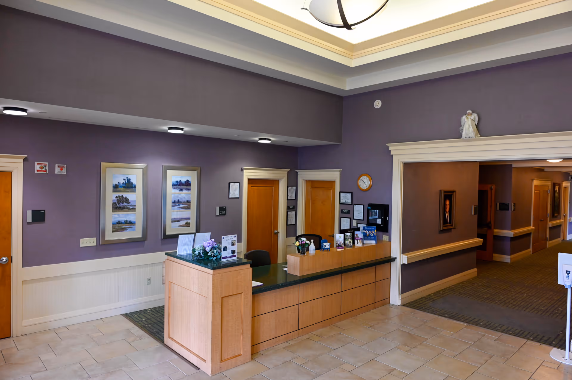 Lobby reception at Alivia Care @ Home with beige desk, purple walls, framed art, and hallway.